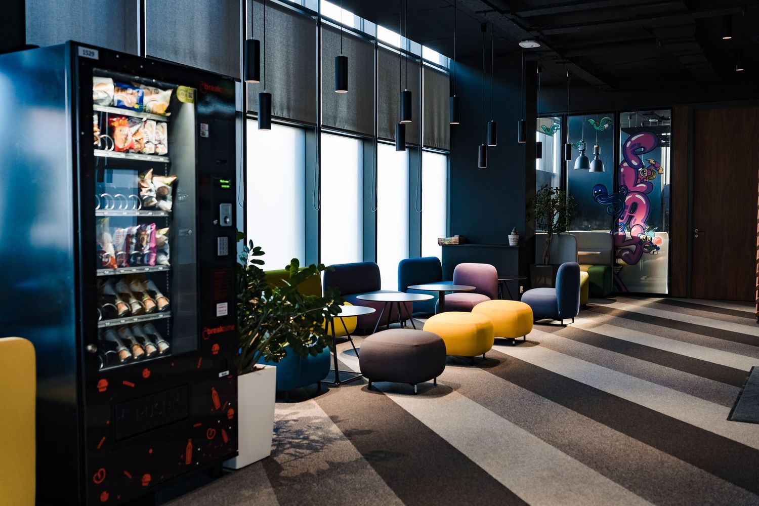 Modern office lounge with a snack vending machine, yellow ottomans, cozy armchairs and large windows.