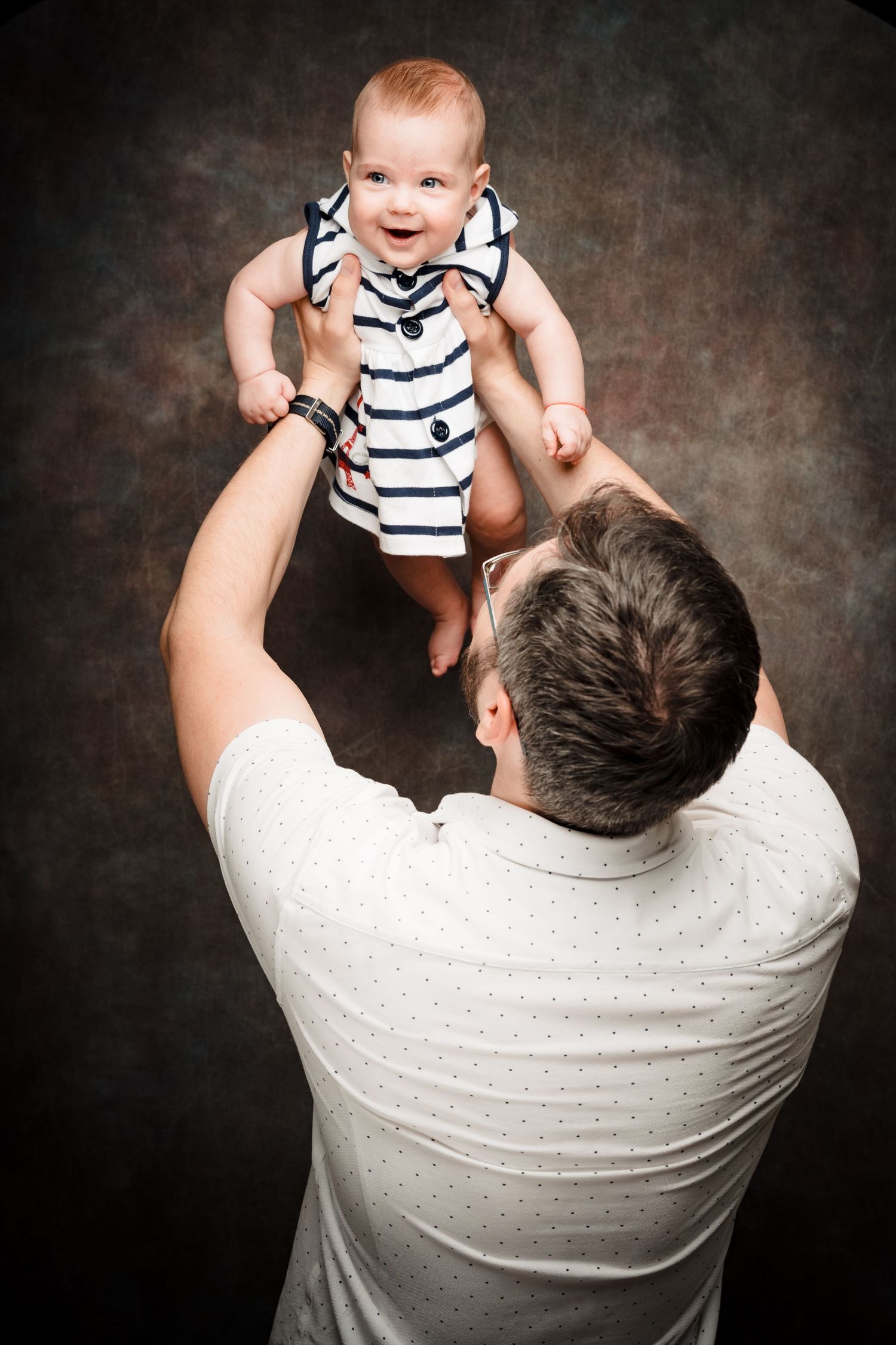 A father in a white polka dot shirt lifts his smiling baby in a striped outfit high above him in a playful studio photo.