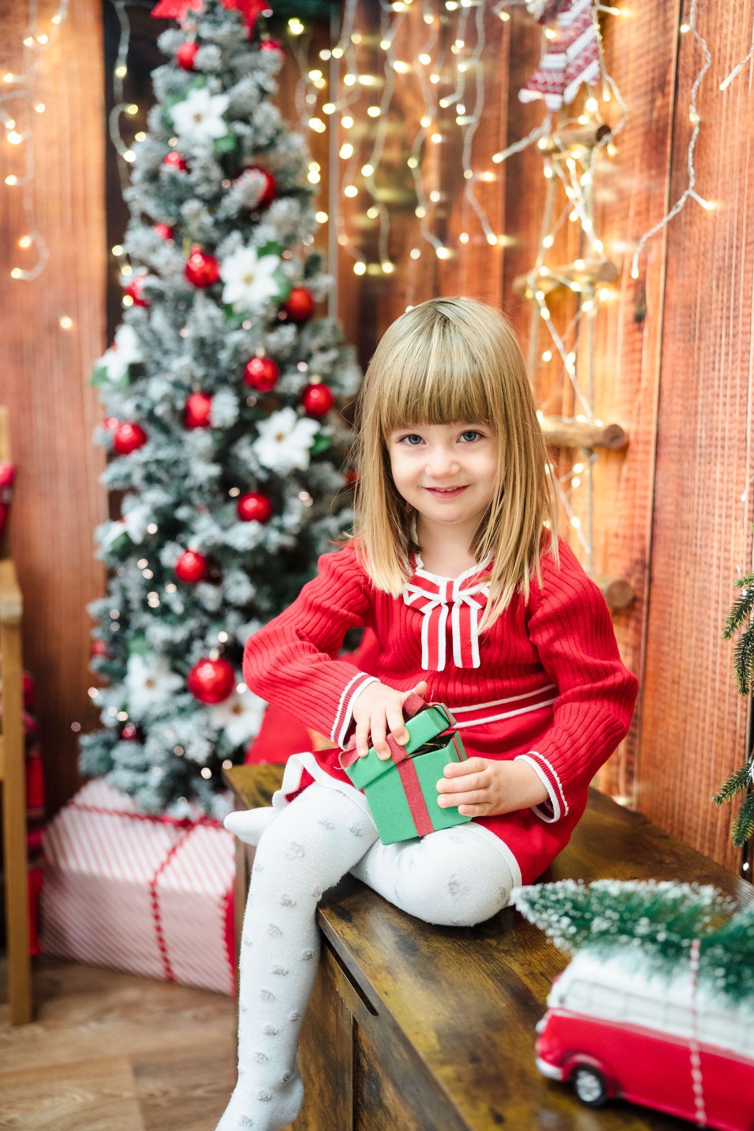 Smiling little girl in a red dress holds a green Christmas gift box beside a decorated tree and festive lights.
