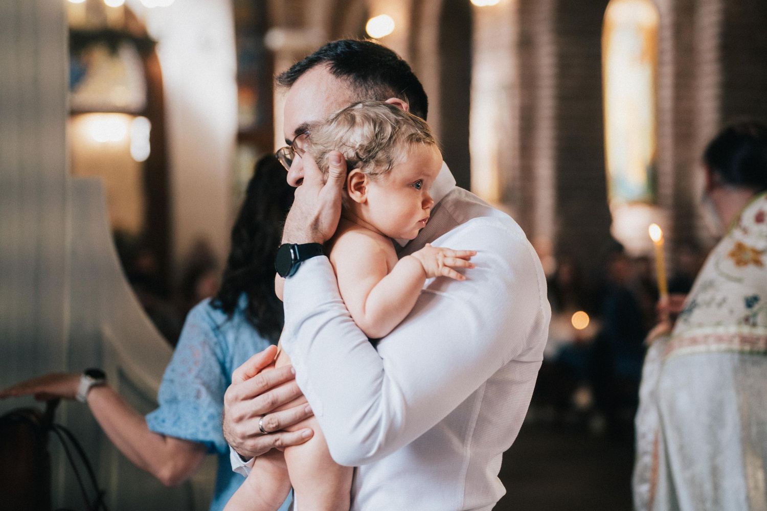 A father in a white shirt tenderly holds his baby close during an Orthodox baptism ceremony inside a softly lit church.