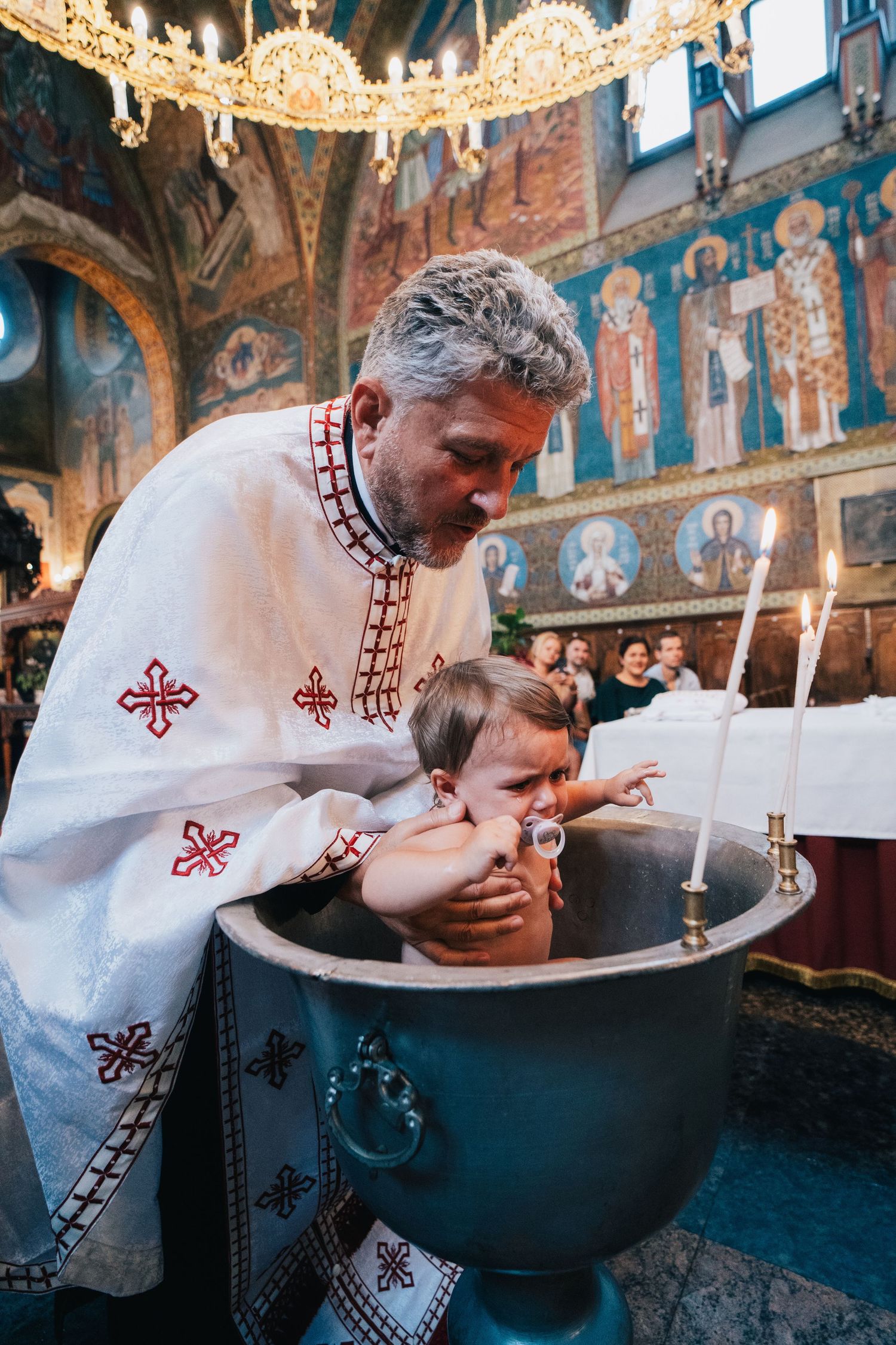 A smiling priest in white vestments baptizes a baby in a metal font as lit candles glow in a church adorned with religious frescoes.