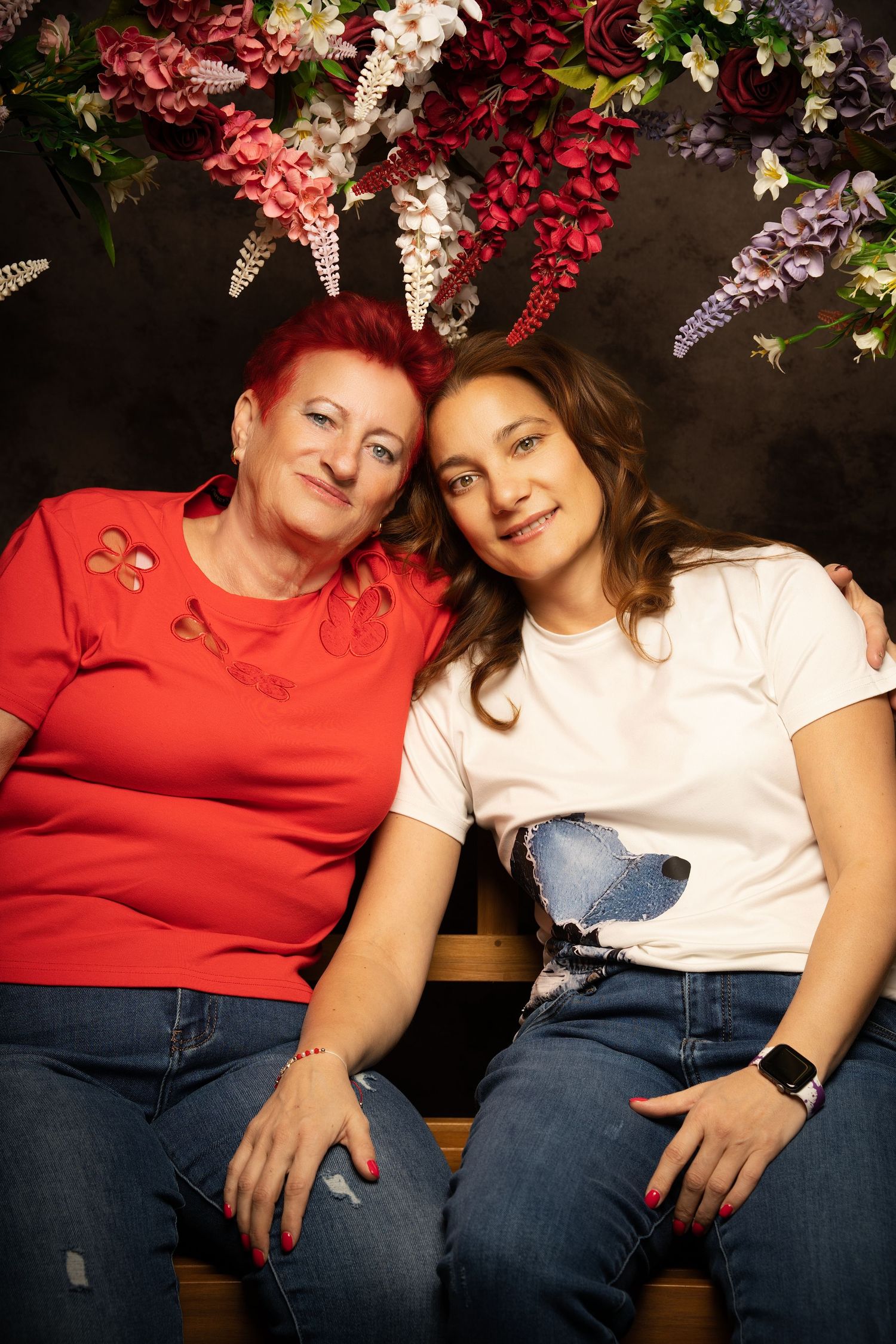 A red-haired woman in a red top and a long-haired woman in a white tee sit close together beneath hanging flowers in a studio.