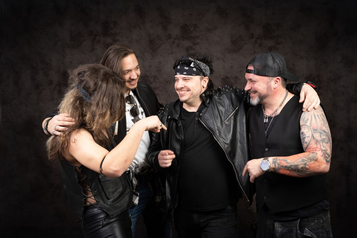 Four rock band members laugh and embrace in a candid group moment wearing leather, bandanas, and tattoos against a dark backdrop.