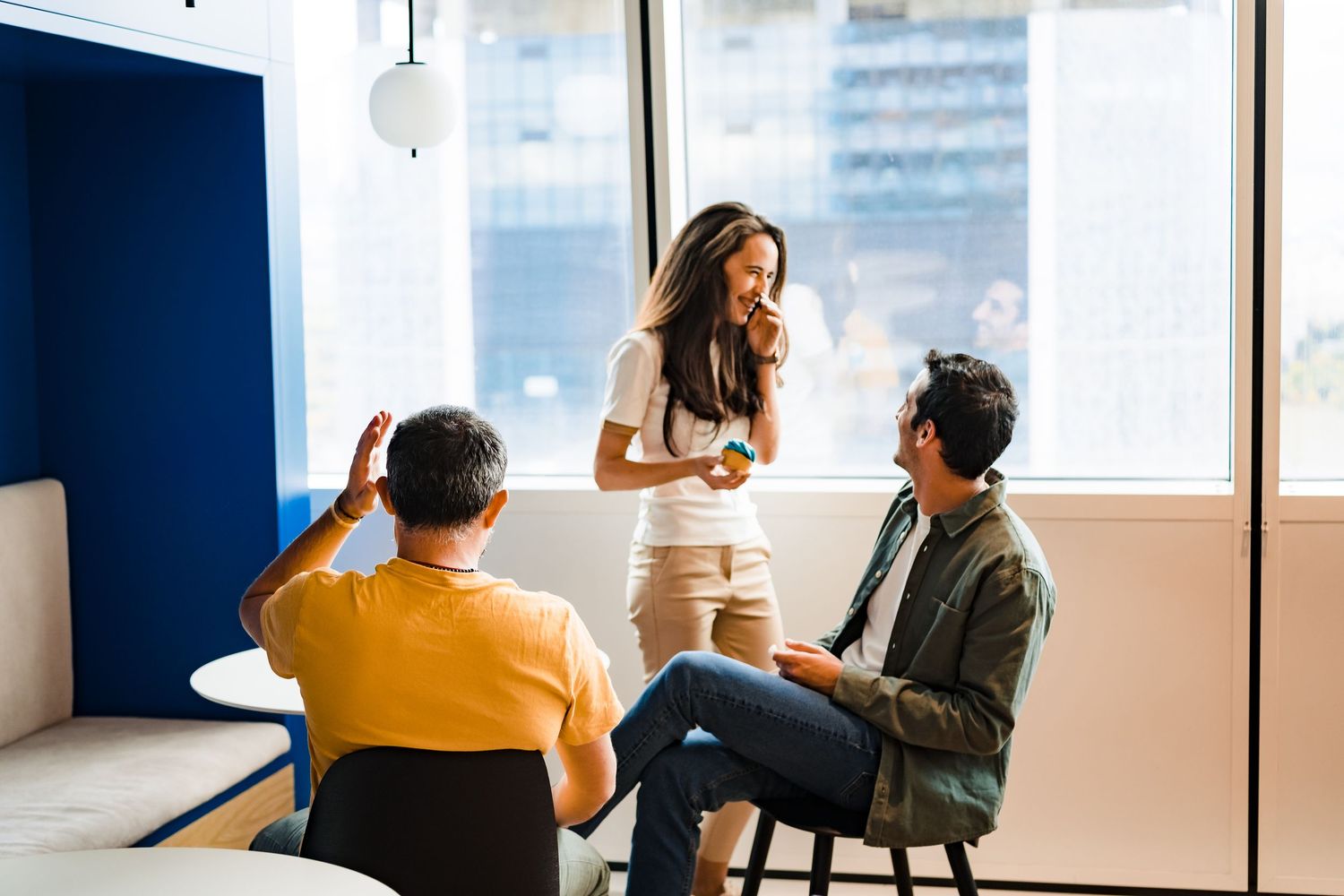 A woman holding a drink stands by a large office window chatting with two seated male colleagues in a casual meeting area.