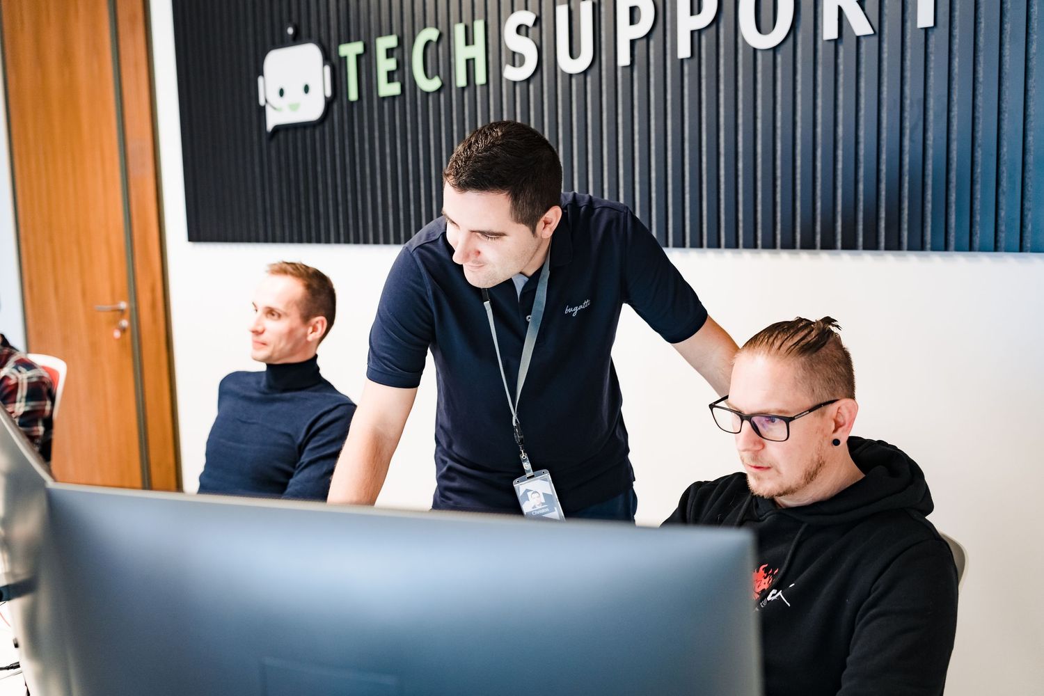 Tech support manager with a lanyard leans over a desk to assist a colleague at a computer in the office.
