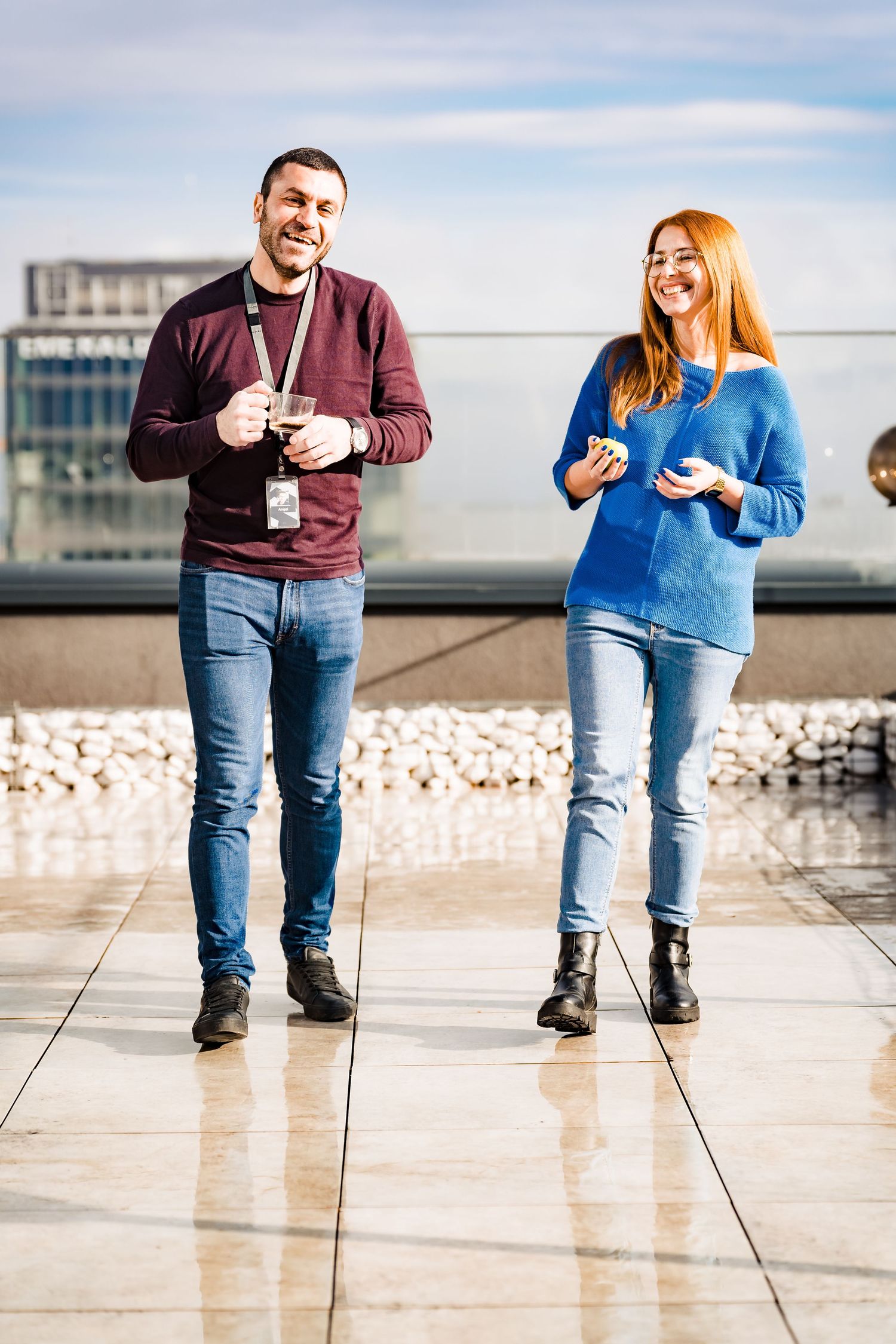 Smiling man with a lanyard and a red-haired woman in a blue sweater walk across a sunny rooftop terrace.