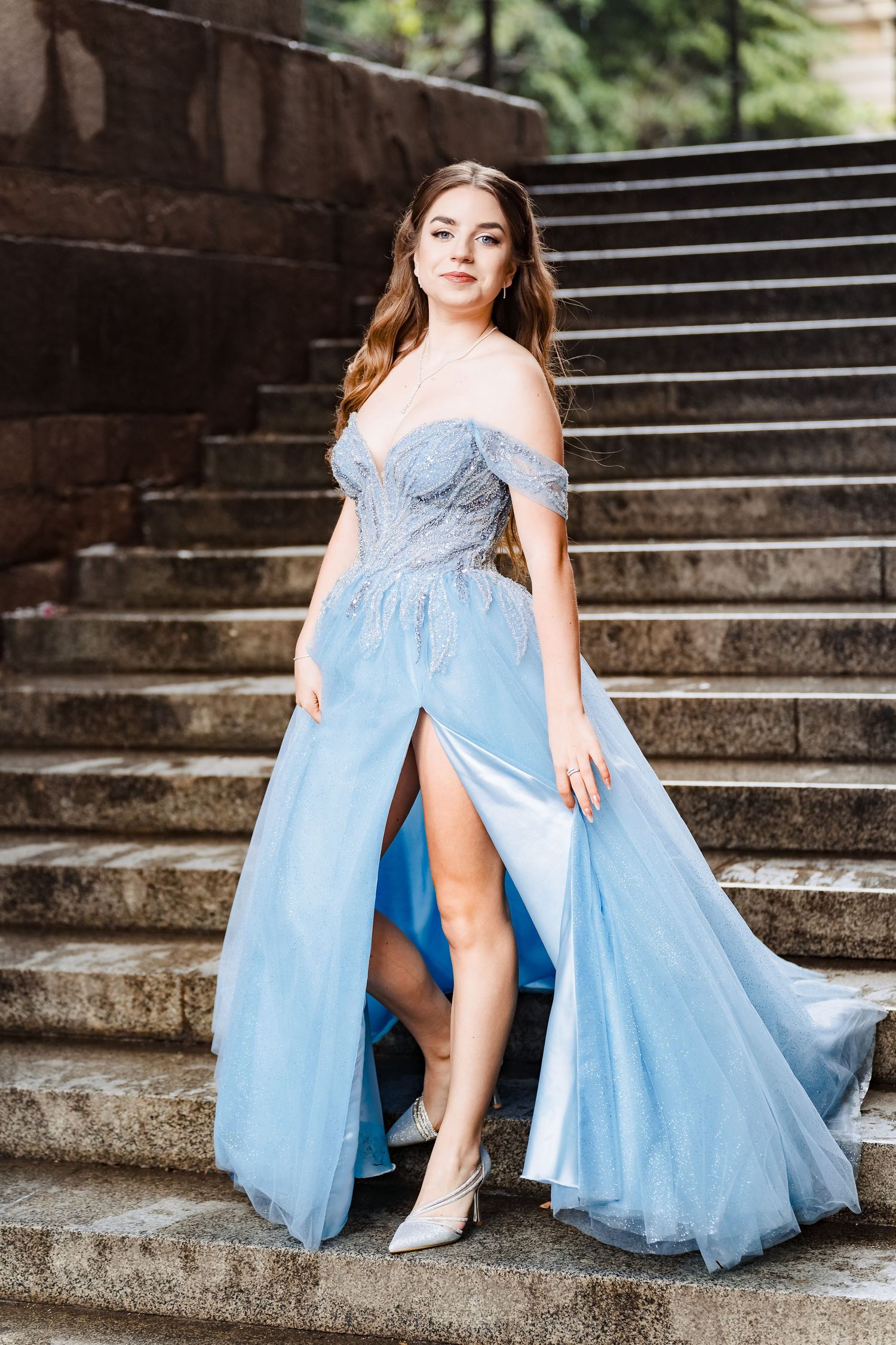 Smiling young woman in a sparkling blue off-the-shoulder prom dress with a thigh-high slit stands on outdoor stone steps.