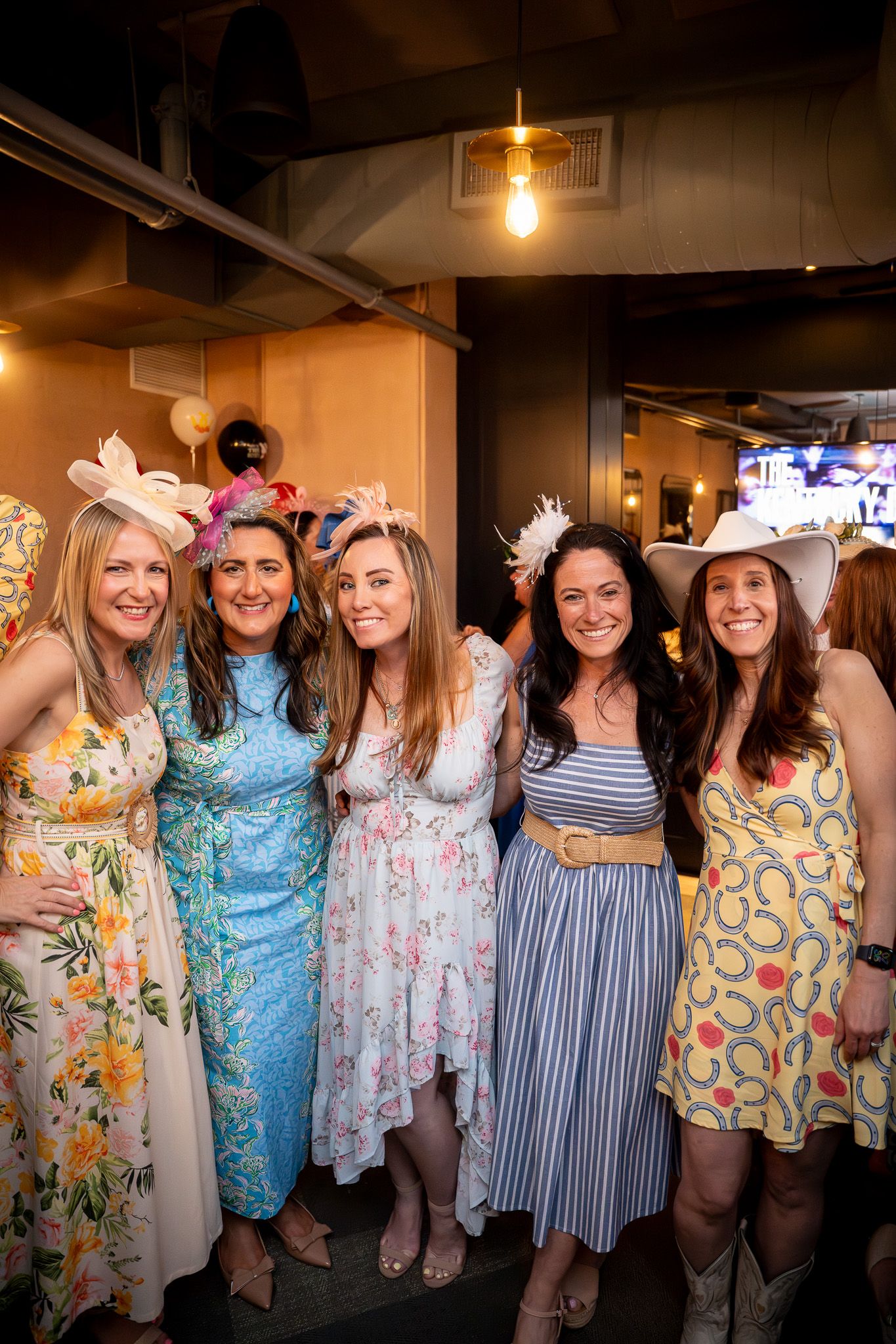 Event photography of a group of well dressed women wearing flamboyant fascinators in front of a step and repeat for a Kentucky Derby party