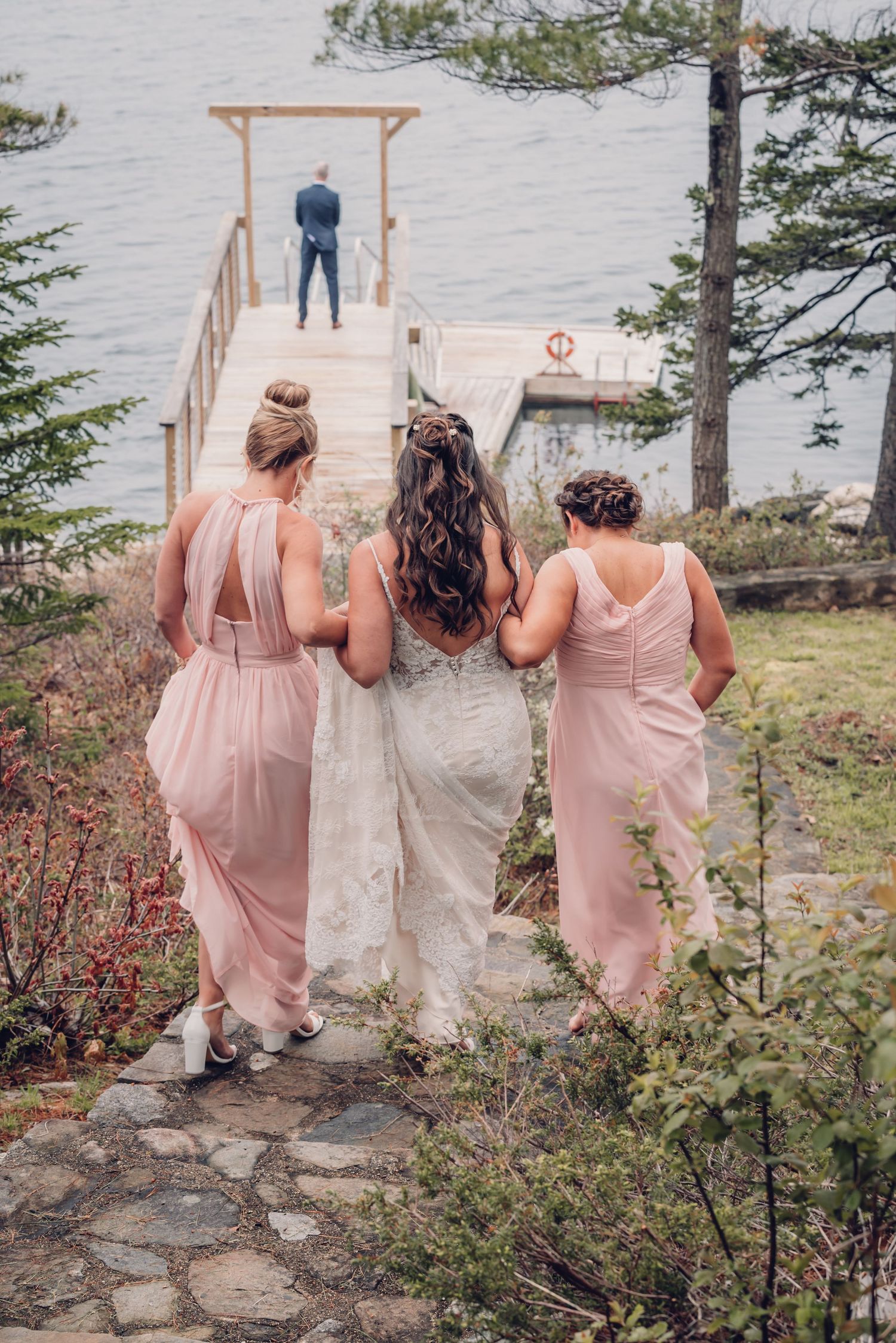 Bride is escorted down a hill by two bridesmaids for first look images with her groom before their lake wedding in New Hampshire