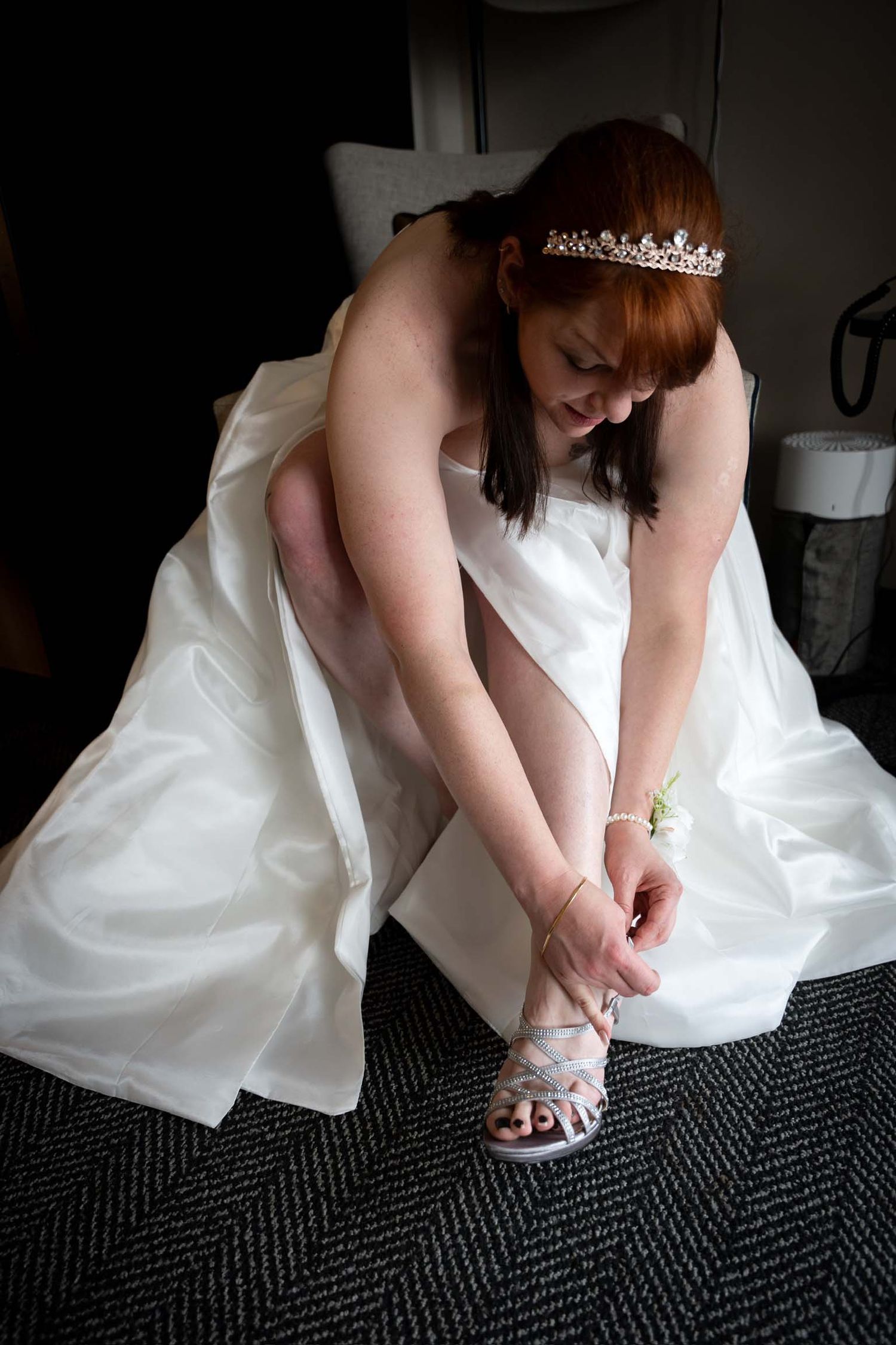 Bride in a classic white satin gown bends to tie her elegant heeled shoes before her glamorous city wedding in the Old Port, Portland, Maine