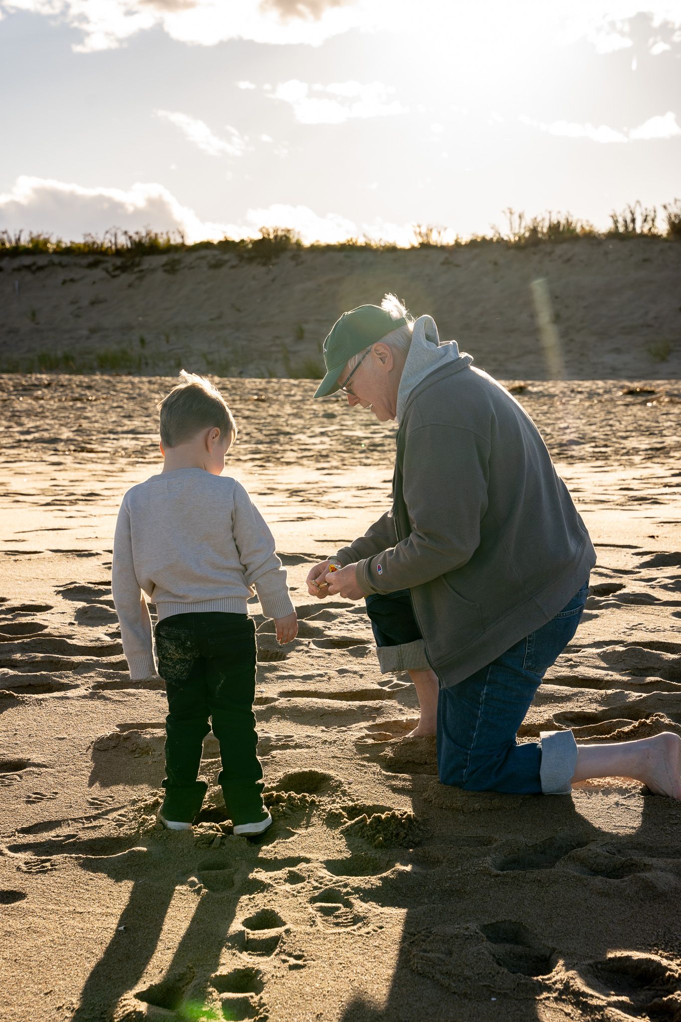 Family portrait of a grandfather picking up a shell on a beach in Newburyport, Massachusetts and offering it to his very young grandson