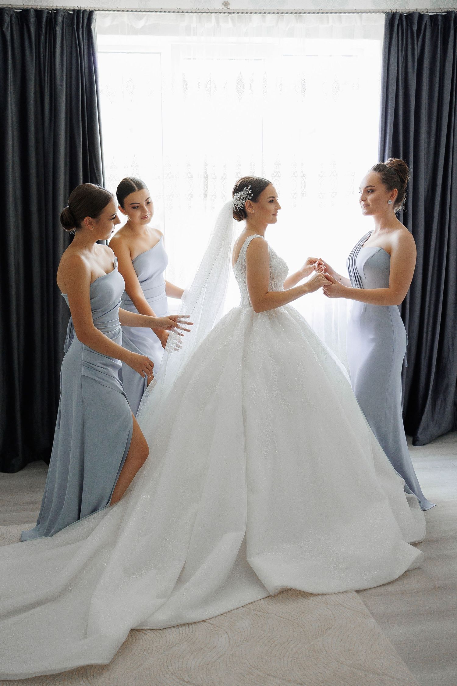 Bride in classic large skirt gown and veil surrounded by her bridesmaids in front of a window before her elegant coastal New England wedding
