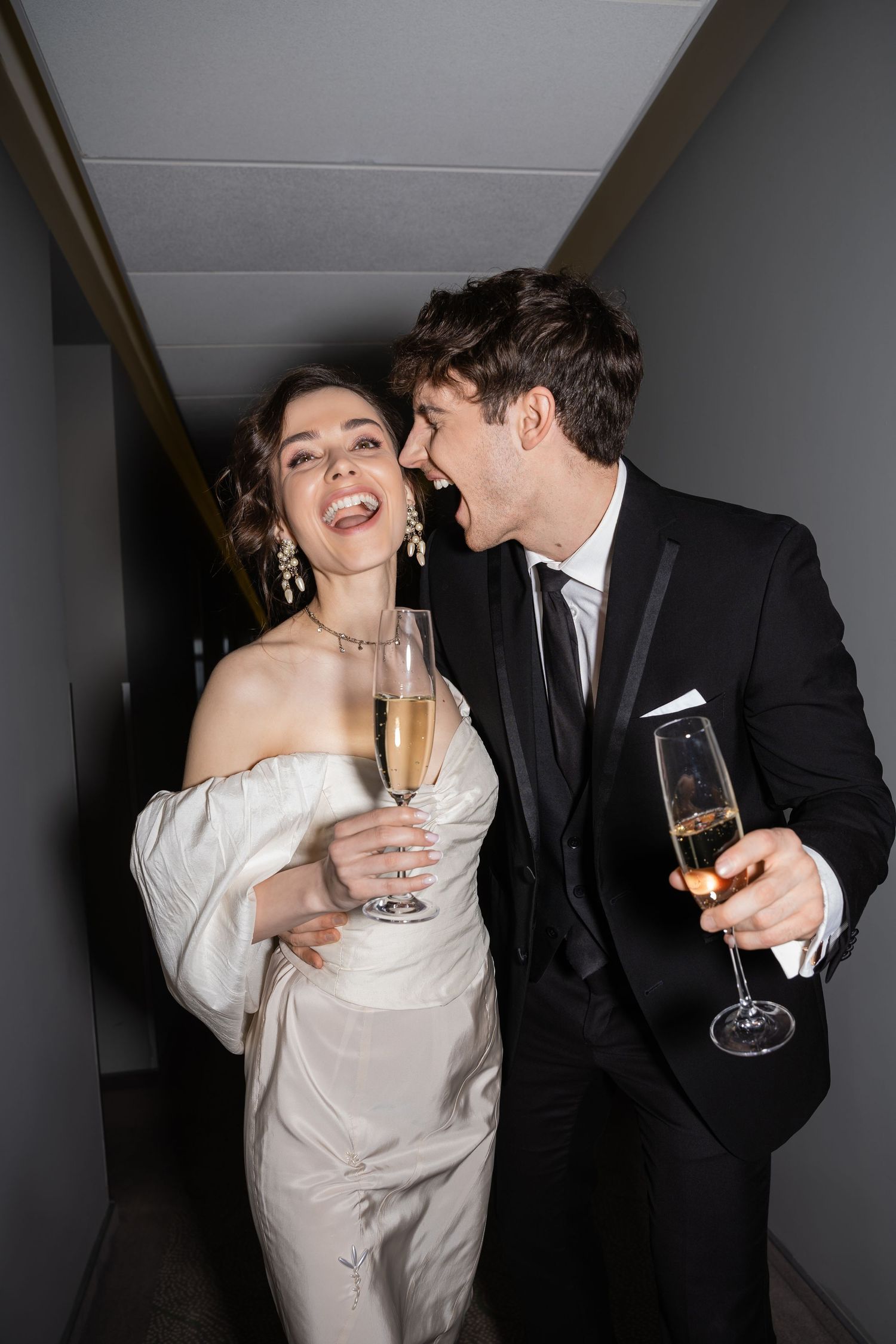 Bride and groom laughing loudly holding glasses of champagne in a hotel hallway after their urban luxury wedding in Boston, Massachusetts