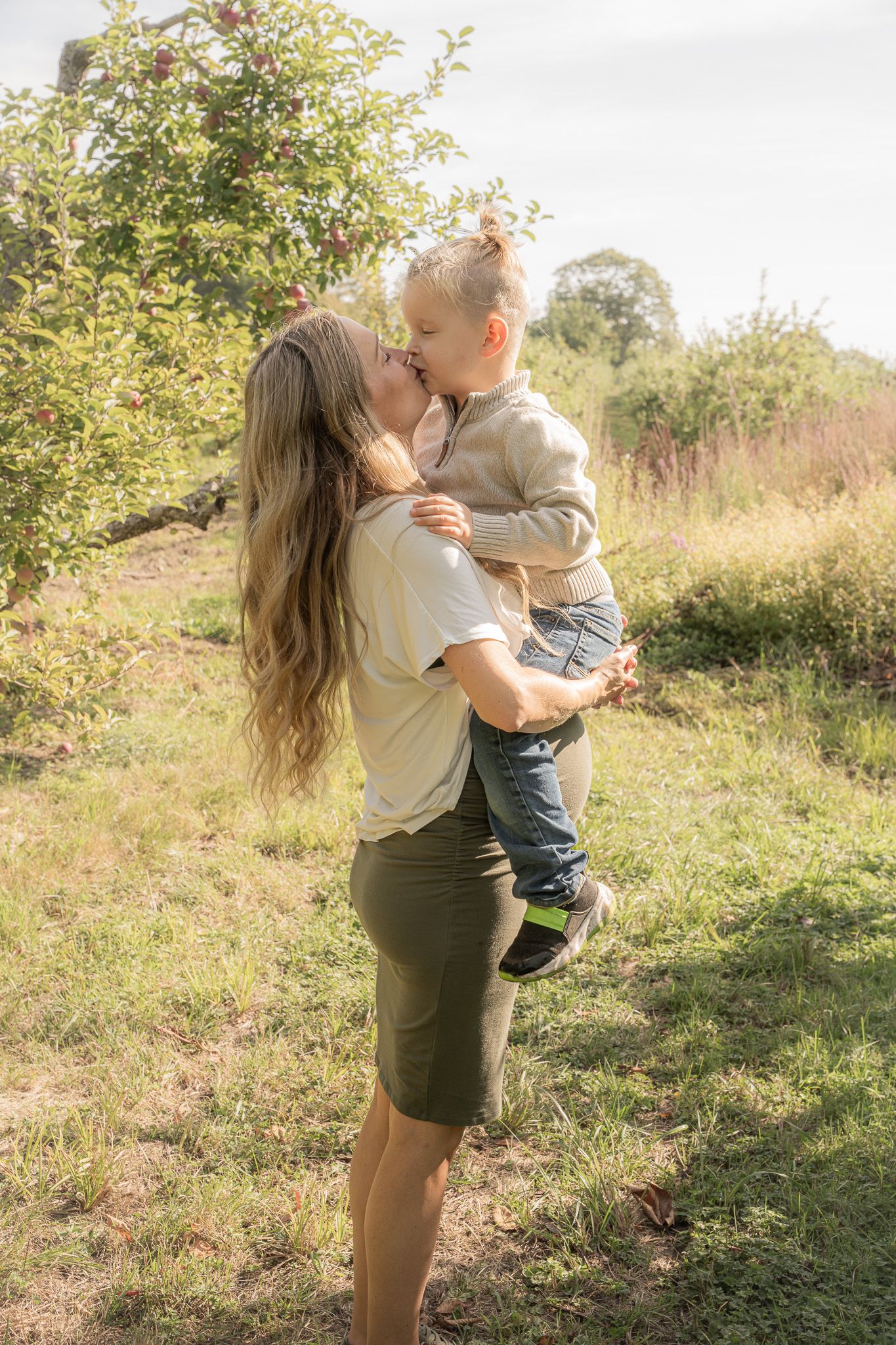 Family portrait of a pregnant mom holding her other young son and giving him a kiss in an apple orchard in Hampton Falls, New Hampshire