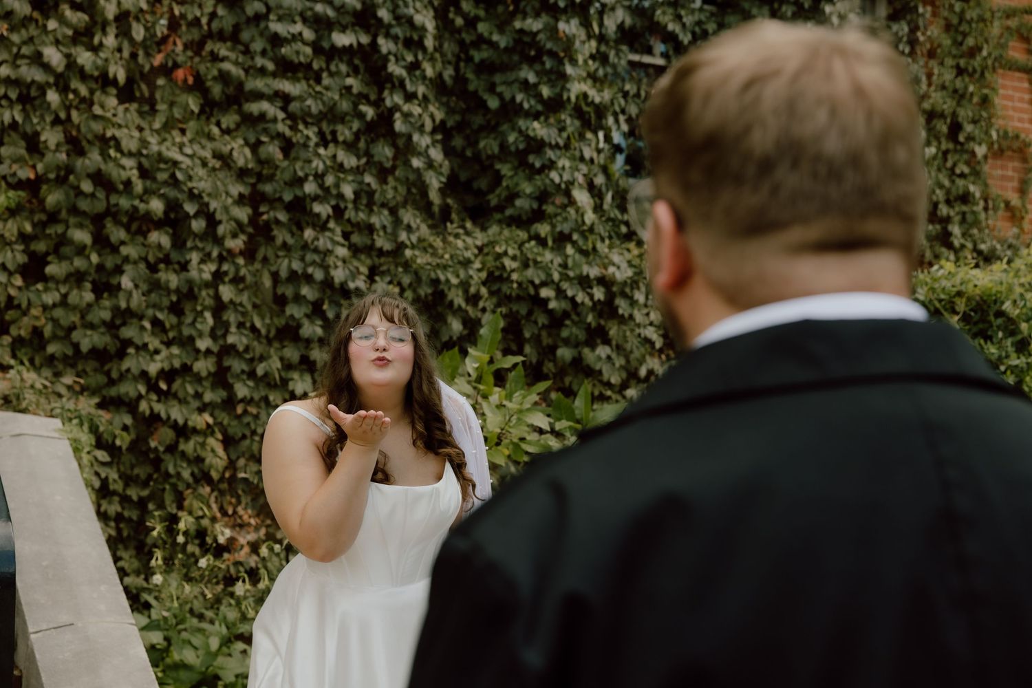 Bride is framed by the back of the grooms head in the editorial style wedding photo. She is blowing a kiss at him for a sweet candid moment.
