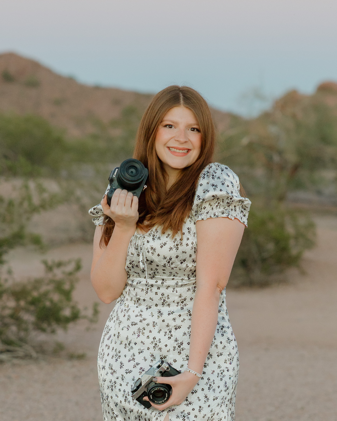 Malachi Westbrook, wedding photographer, is photographed here holding her cameras while in front of Arizona mountains and landscape.
