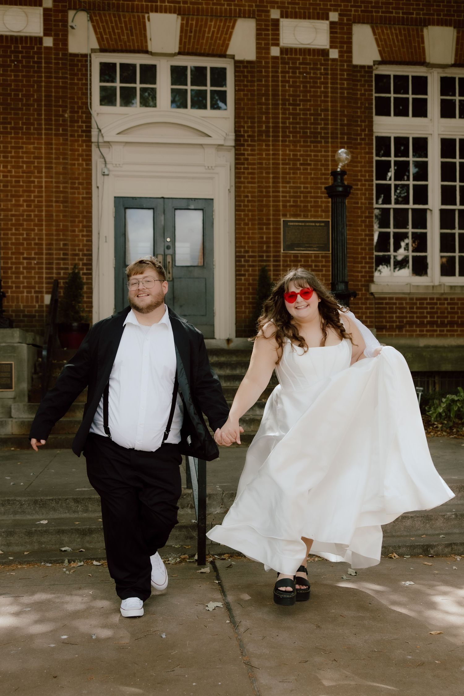 Bride and groom in a fun photo laughing and showing off heart shaped sunglasses in front of downtown Fayetteville building.
