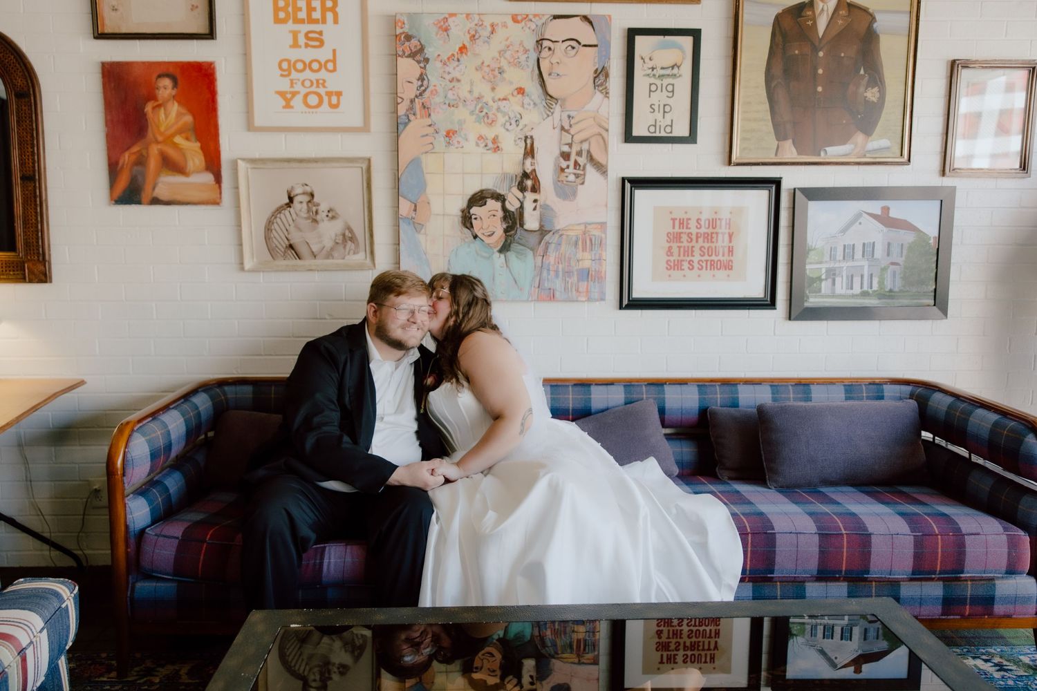 Bride and groom kissing on blue plaid couch in front of gallery art wall at the Graduate Hotel in Fayetteville, Arkansas after eloping.