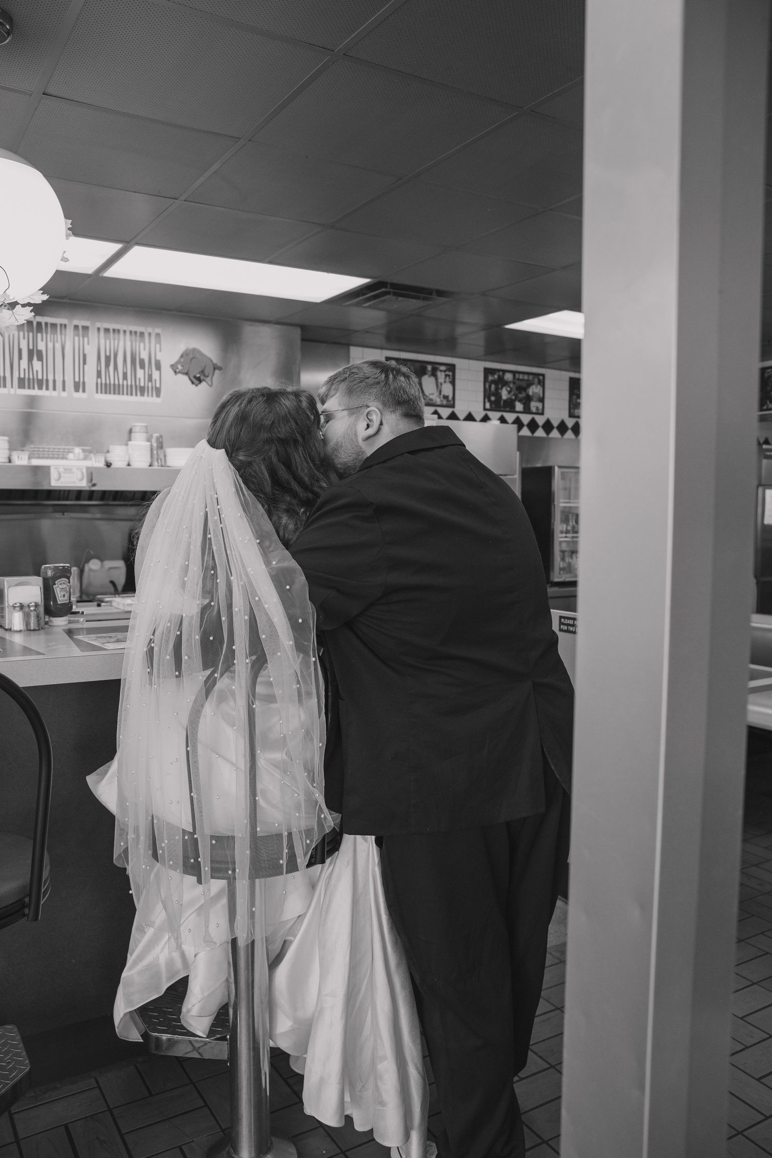 Bride and groom sitting at the bar of the Waffle House caught in a documentary style black and white photo kissing showing brides veil.