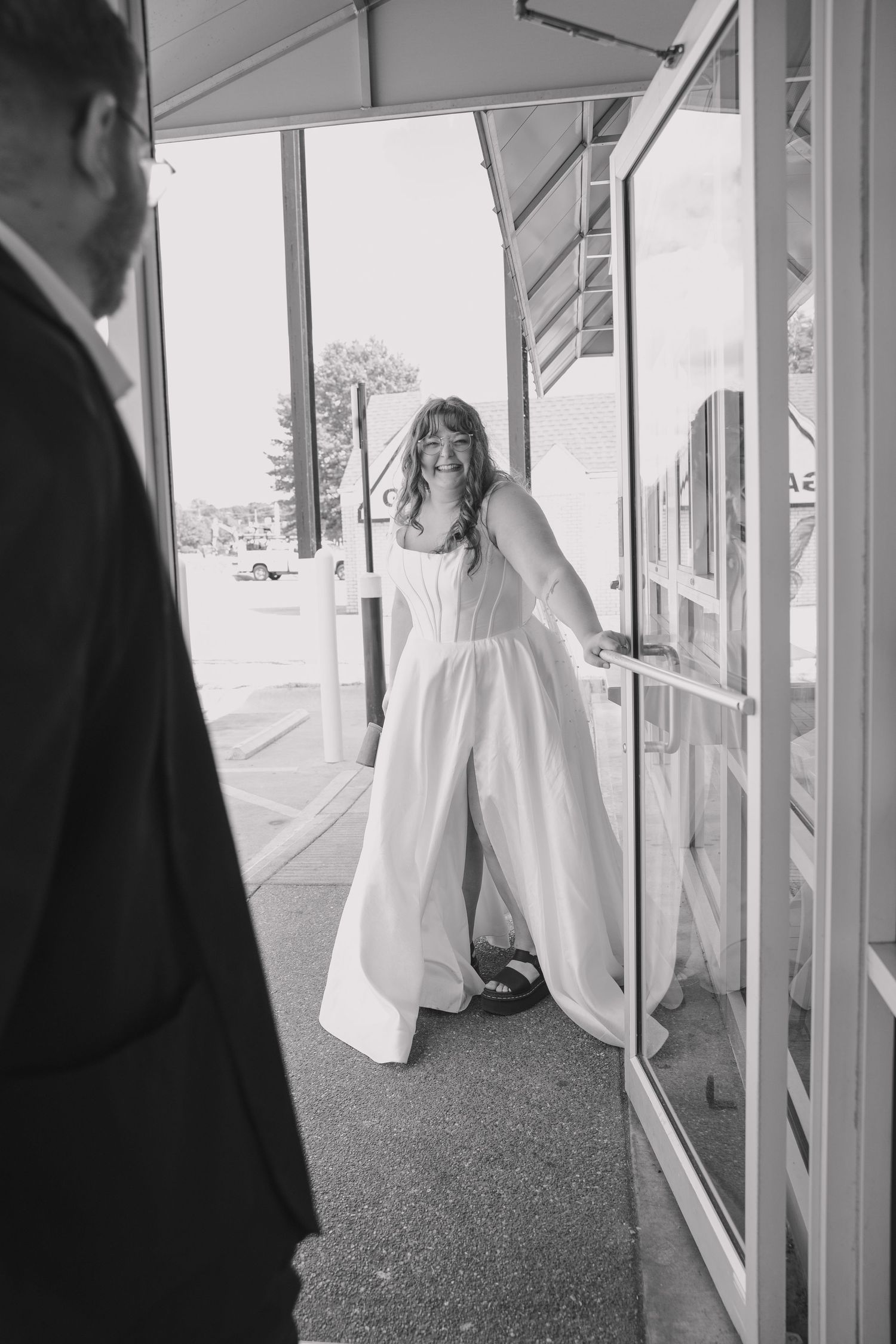 Documentary style photography moment in black and white of bride holding open the door to Waffle House laughing at groom as he exits.
