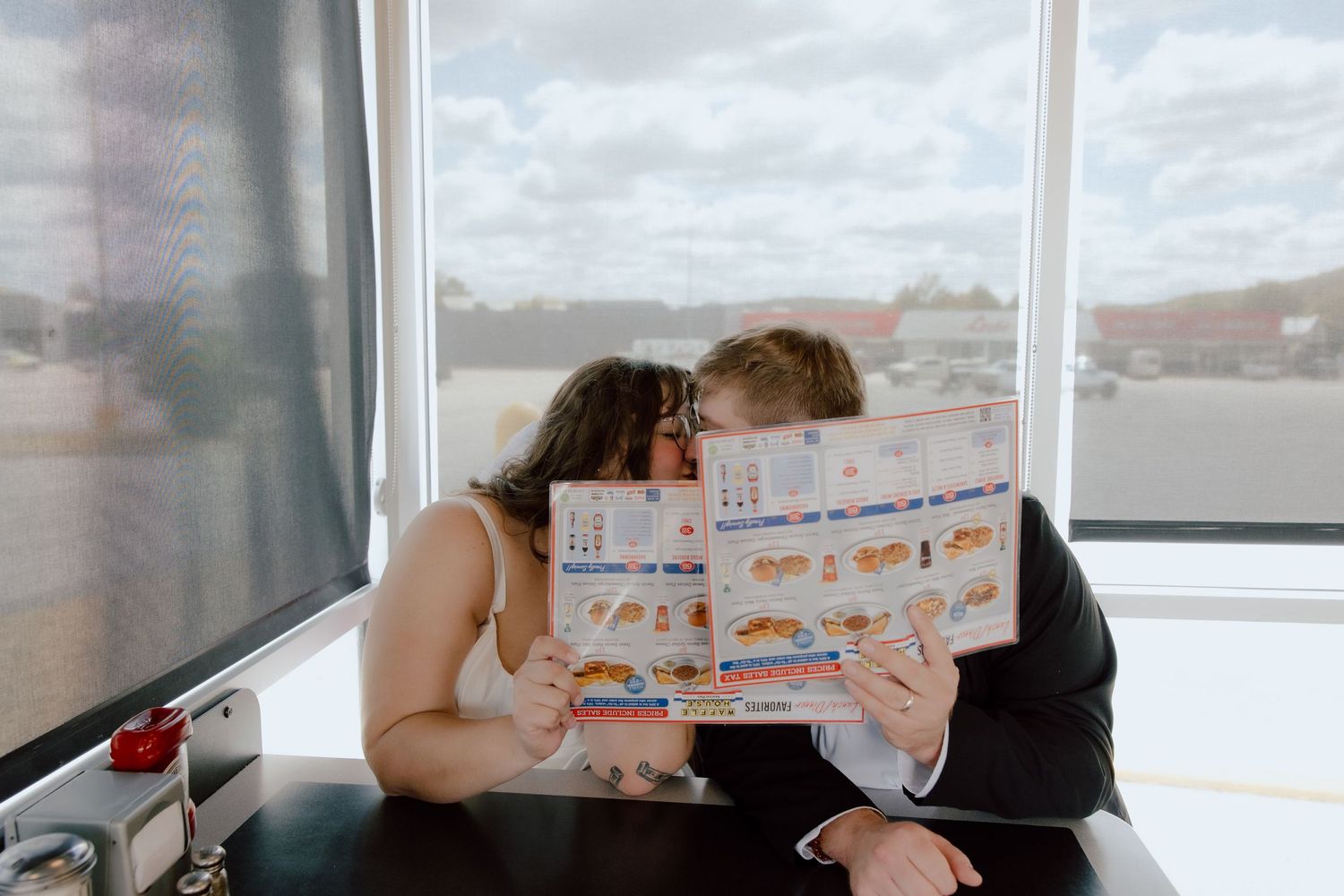 Bride and groom hiding behind Waffle House menus during their post wedding meal, sharing a kiss in their gown and suit.