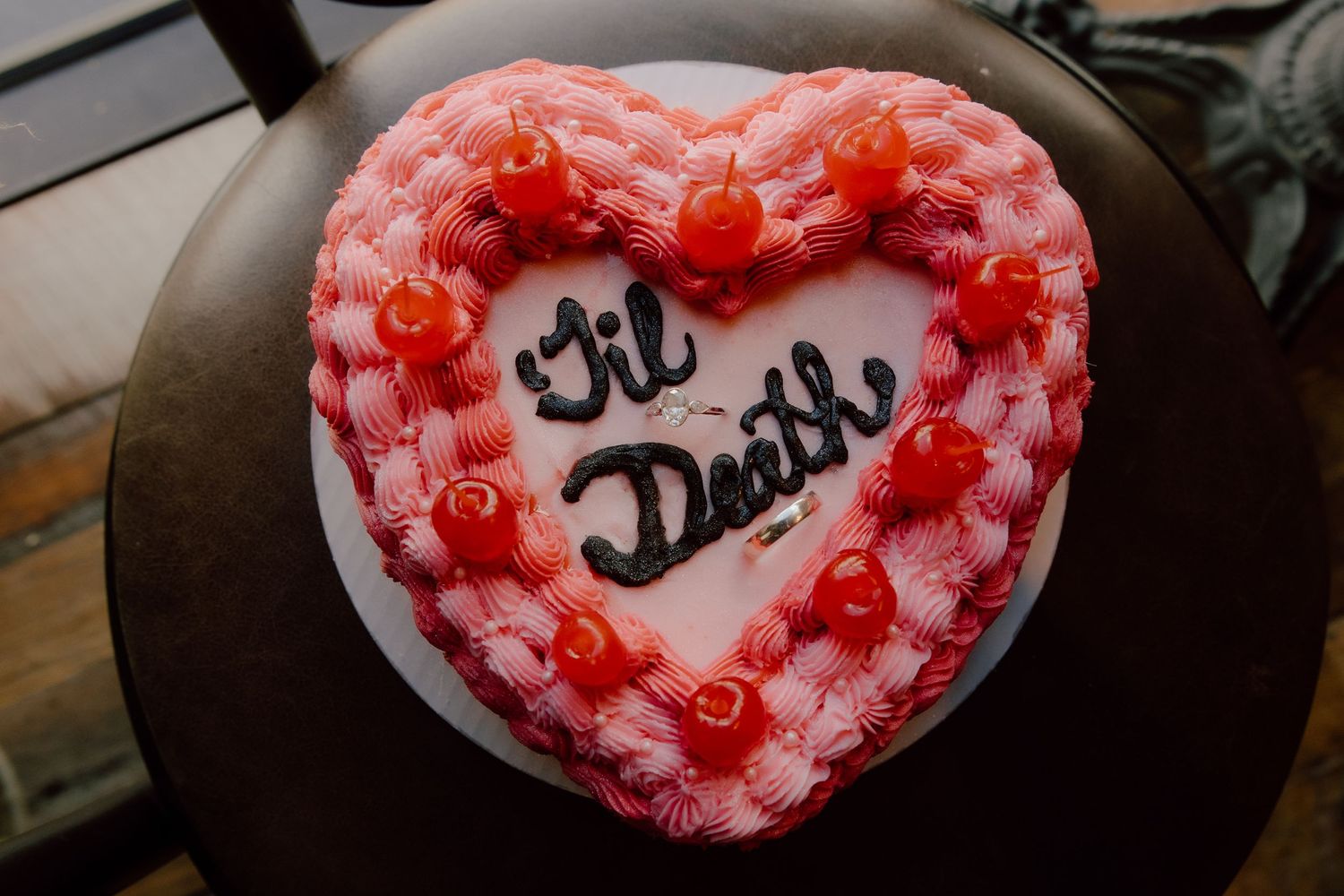 Pink heart shaped wedding cake with 'til death written on it and the couples wedding rings placed inside of the cake with cherries around it