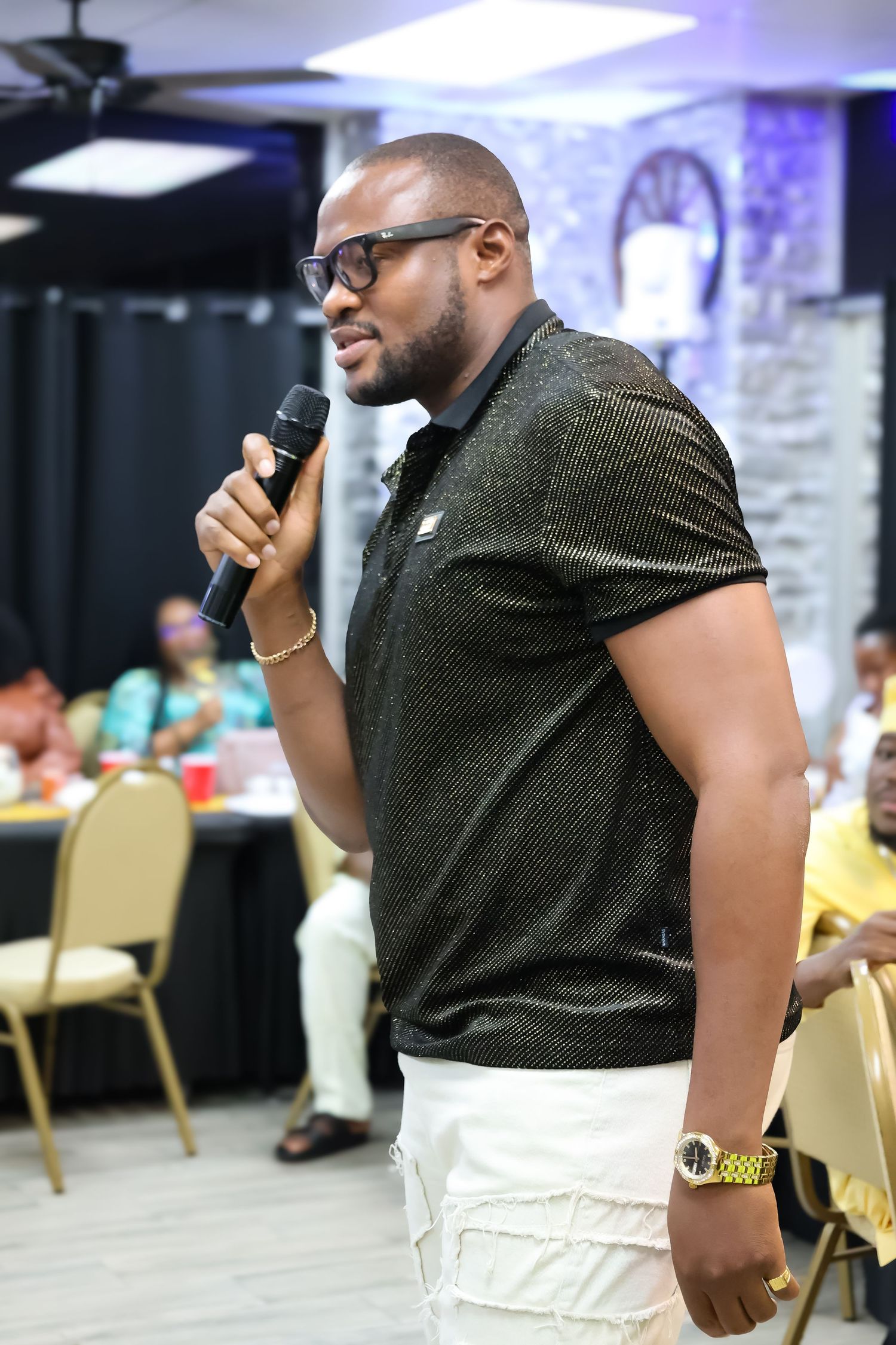 Man in black sparkle polo and white pants speaks into microphone at indoor event.