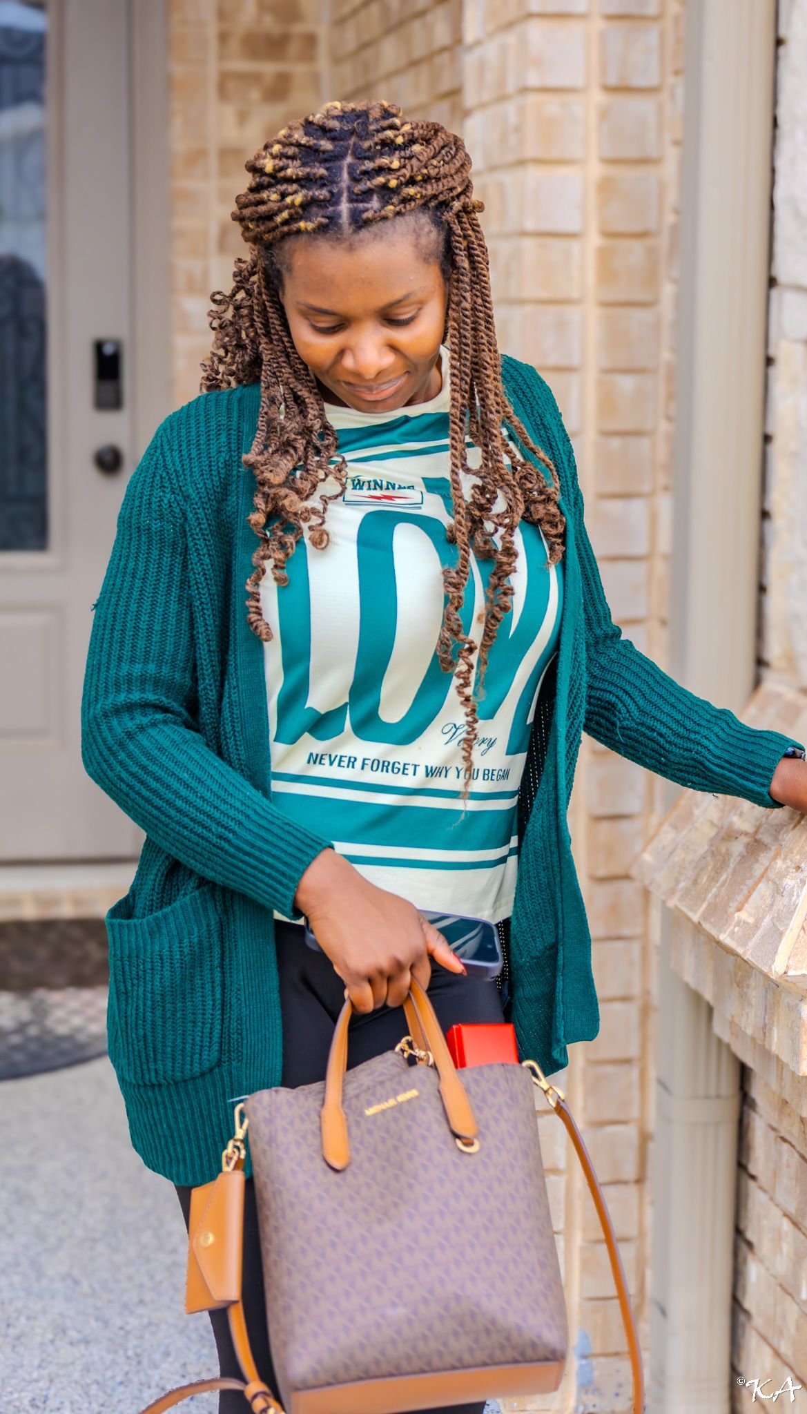 Woman with braided hair wearing teal cardigan and graphic tee, carrying designer handbag near brick building.