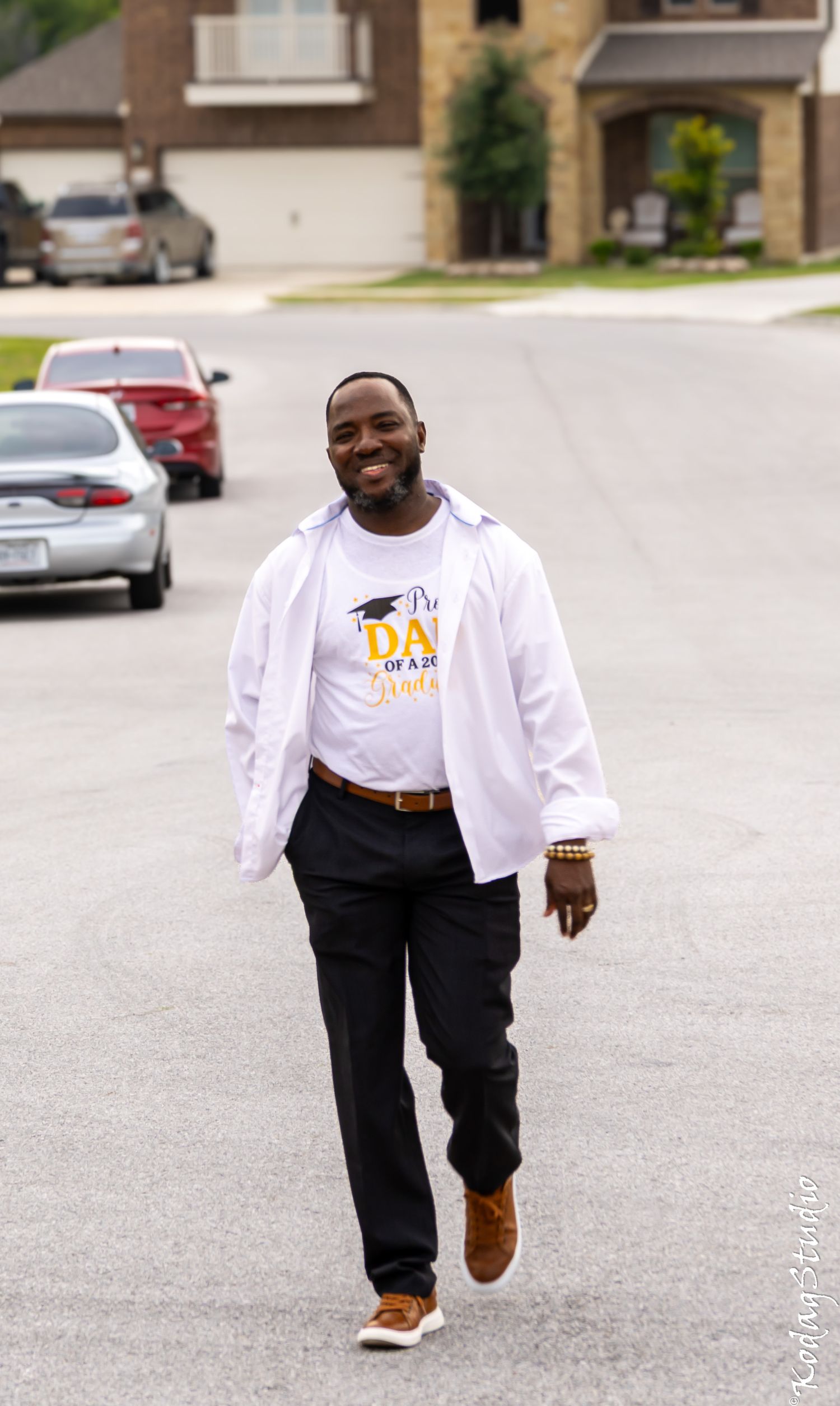 Smiling man walking down a suburban street wearing a white jacket and a Dad of a Graduate t-shirt.