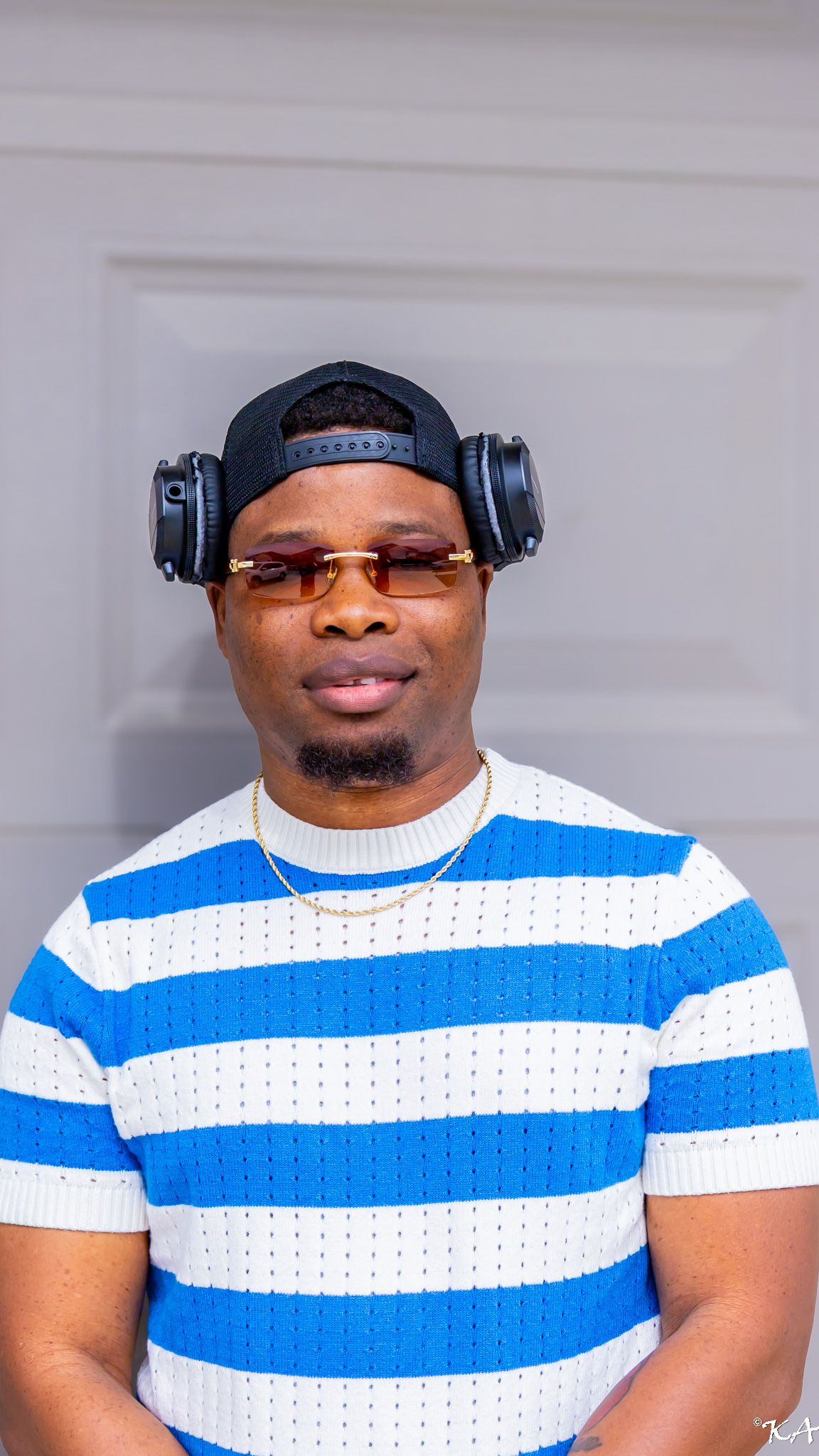 Man wearing blue striped shirt, black snapback, headphones, and gold chain necklace posing outdoors.