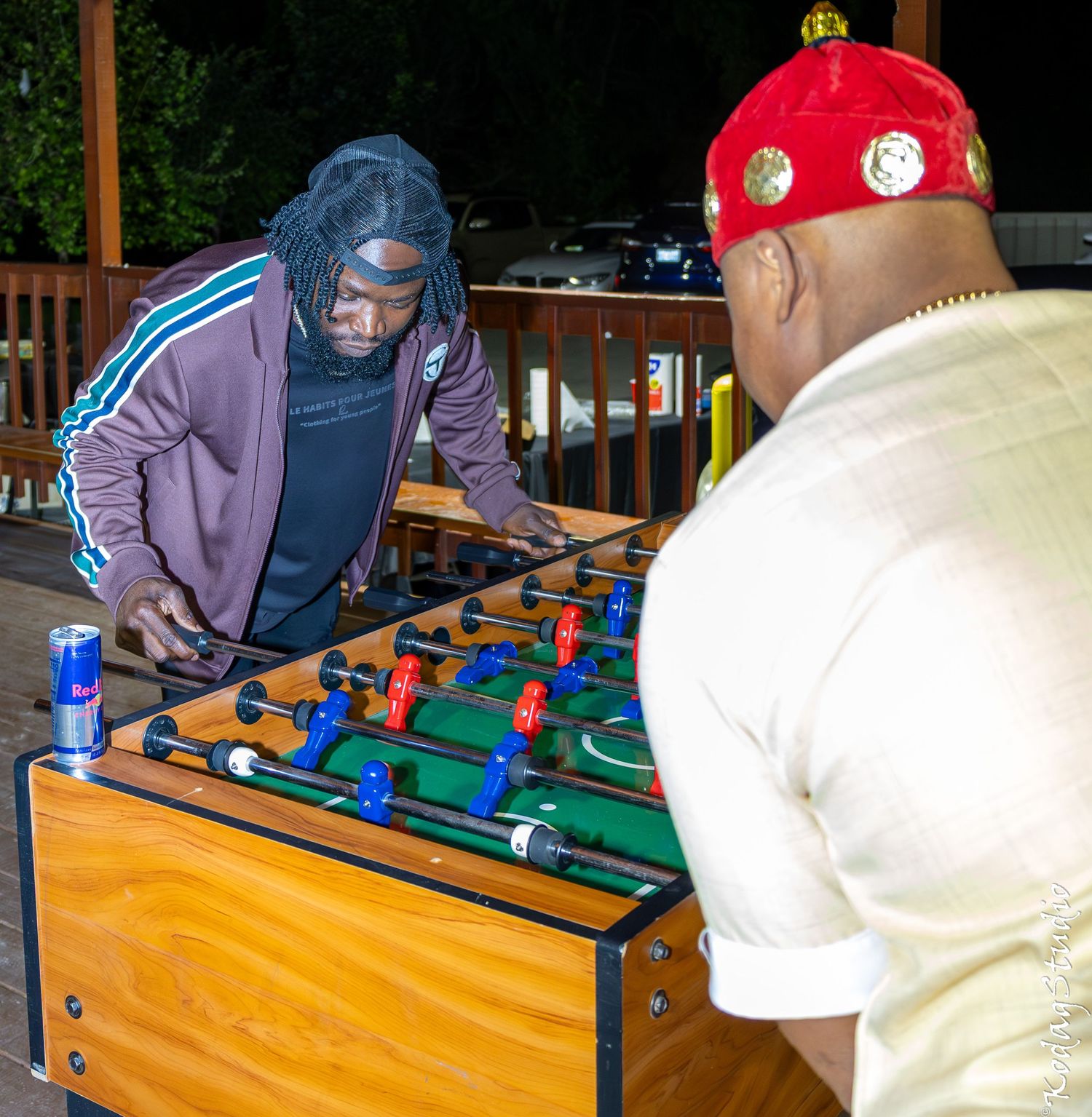 Two men playing foosball outdoors at night, one wearing a red hat, with a Red Bull can on the table.