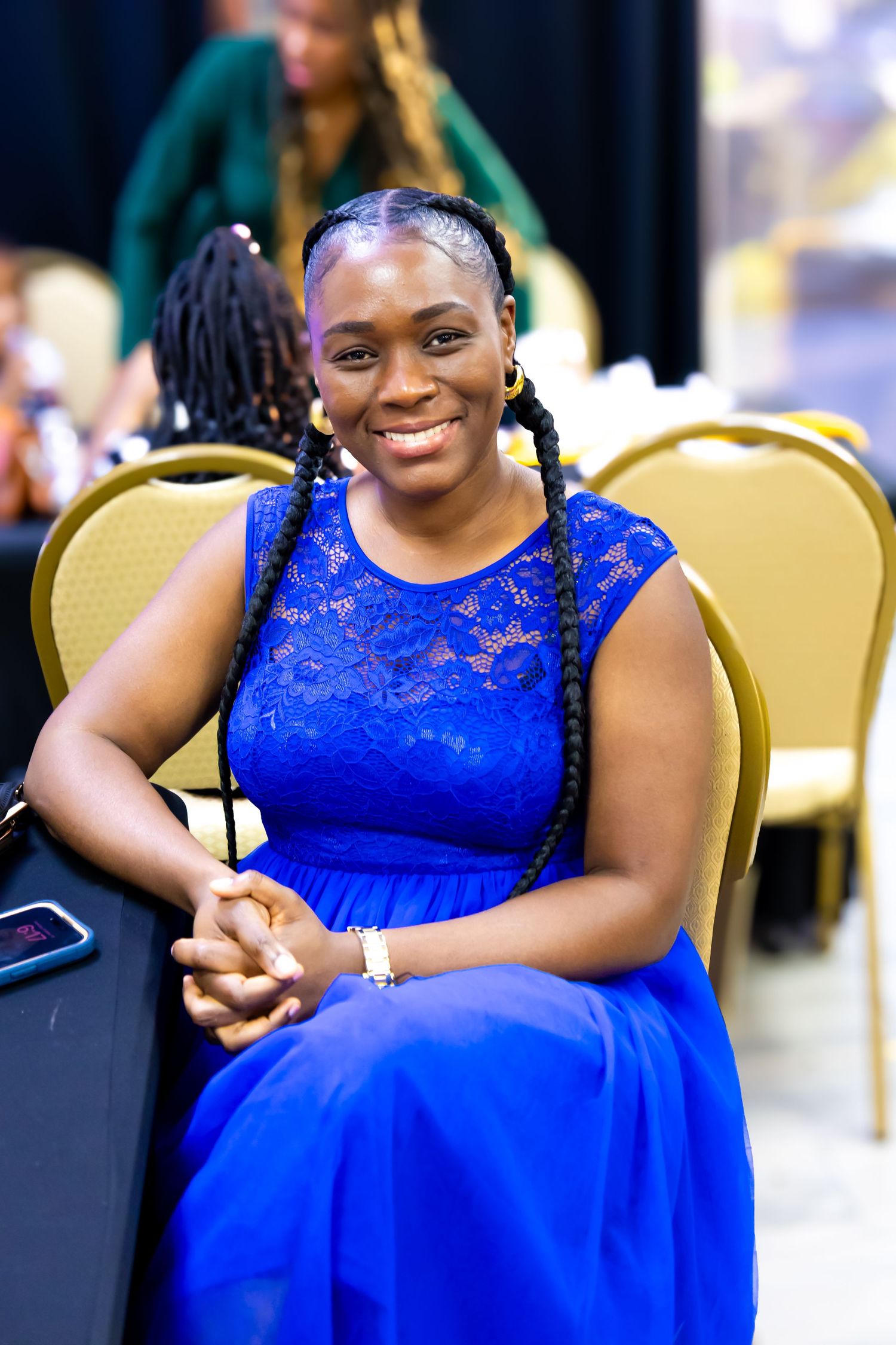 Smiling woman in blue lace dress with braids sits at event table, wearing gold earrings and bracelet.