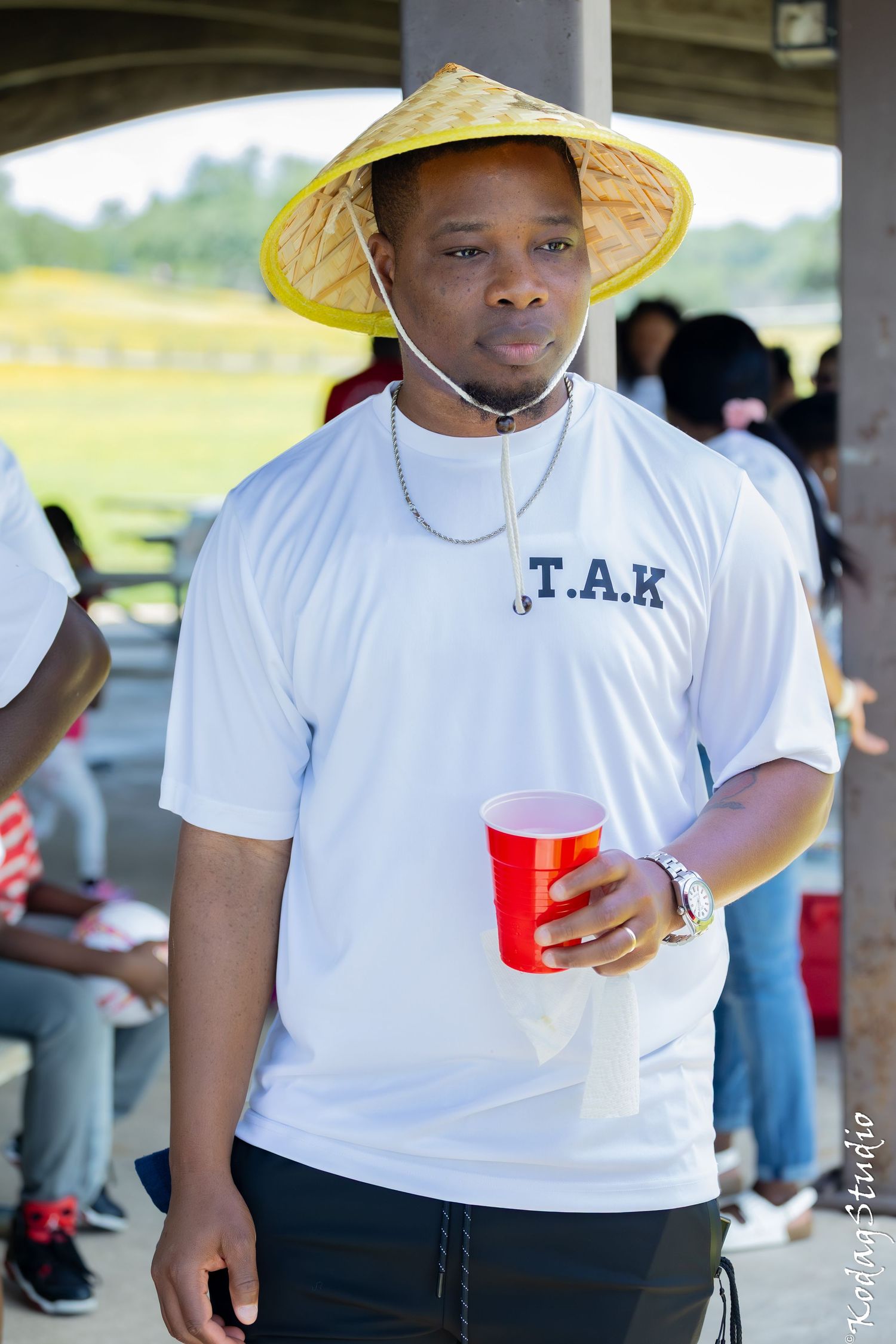 Man wearing a straw hat and white T.A.K t-shirt holding a red cup at an outdoor gathering.