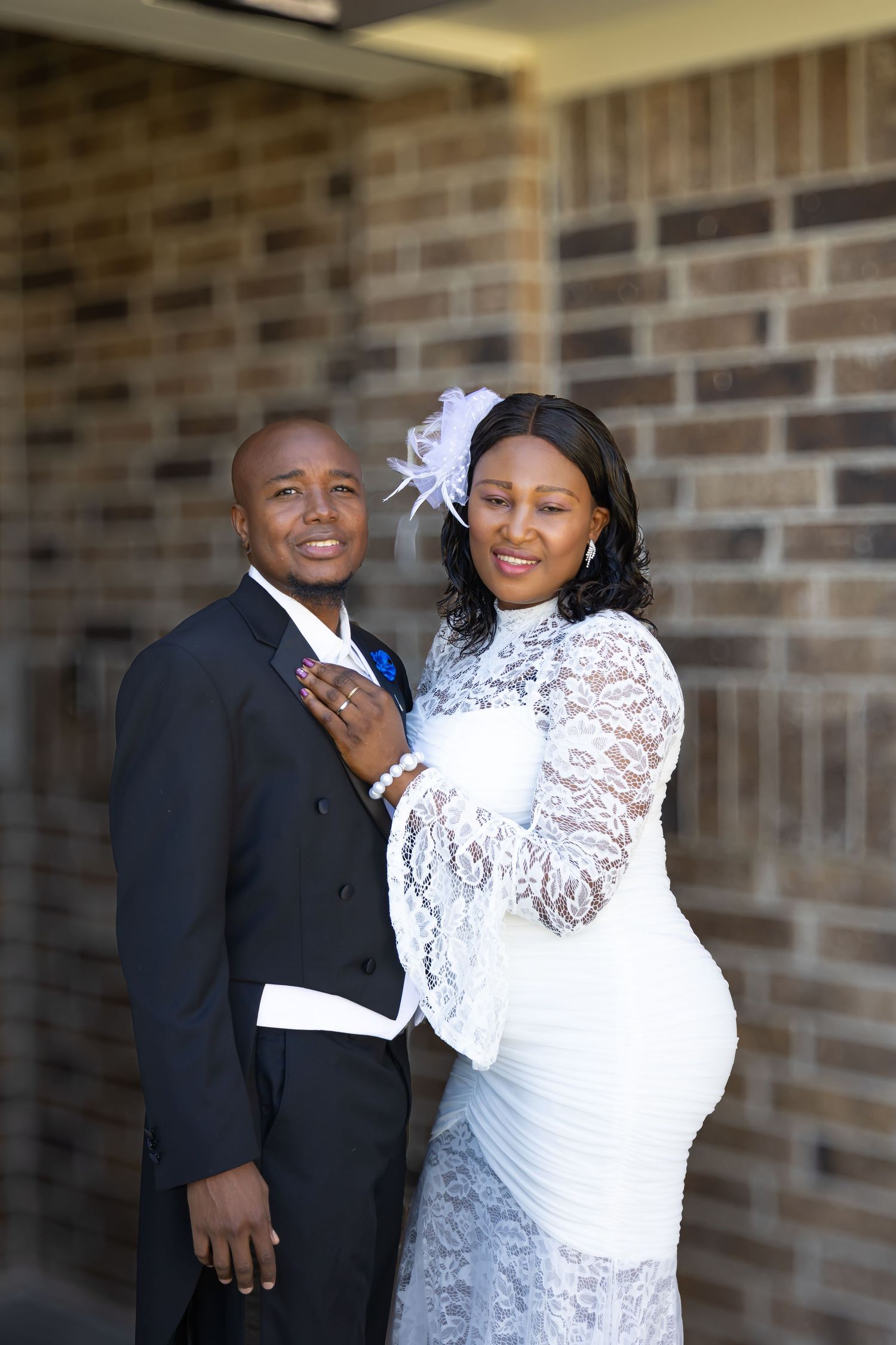 A smiling couple in wedding attire poses before a brick wall, the bride in white lace and groom in a black suit.