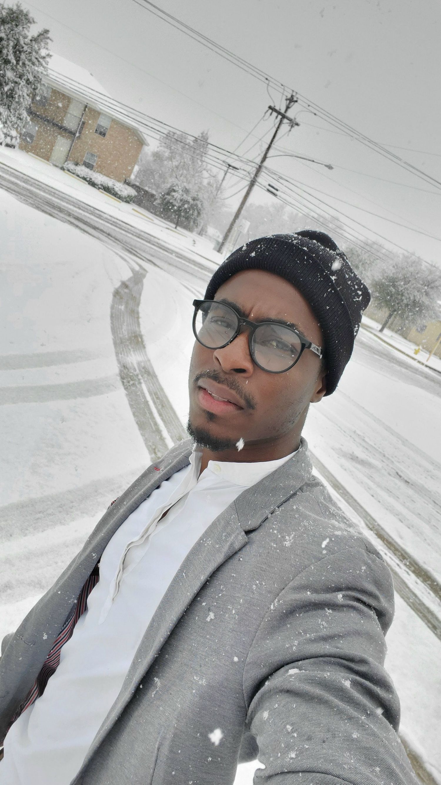 A man wearing glasses and a beanie takes a selfie outdoors during a heavy snowfall on a snow-covered street.