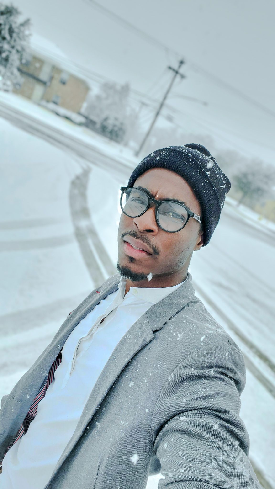 Man in gray blazer and beanie taking a selfie outdoors in snowy winter weather conditions.