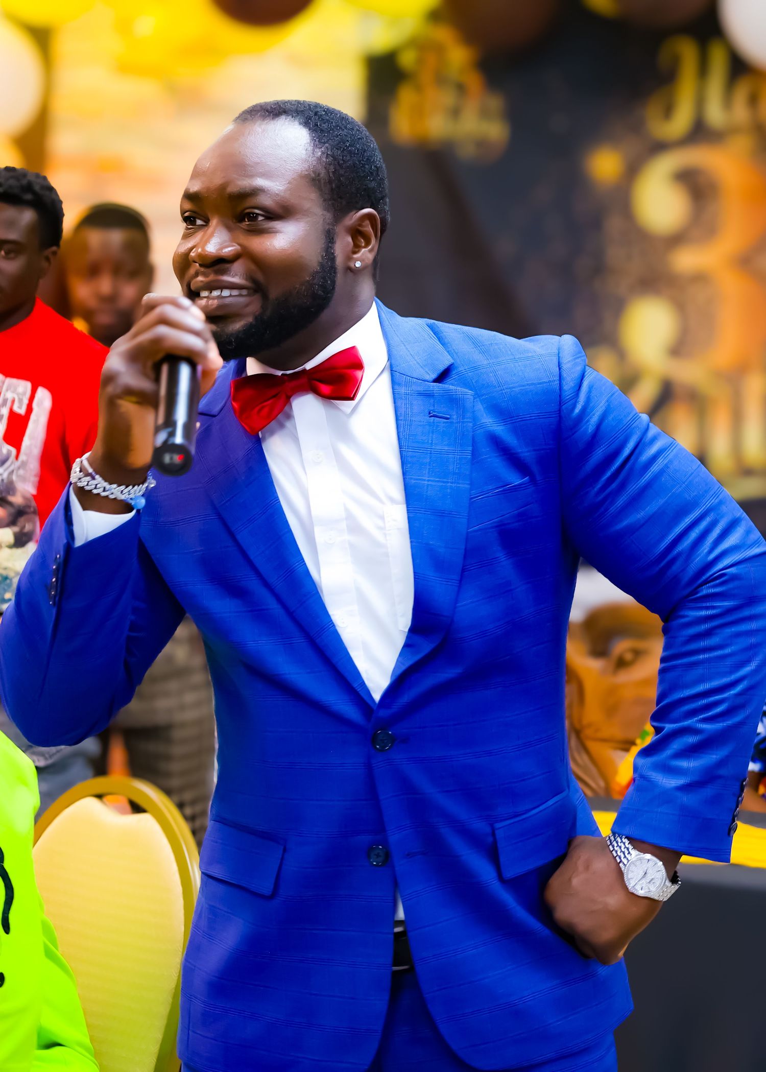 Man in blue suit and red bow tie speaking into microphone at celebration event.