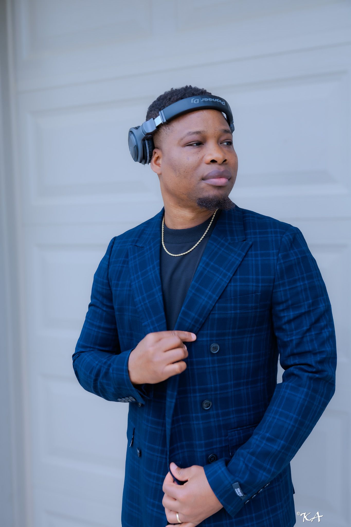 Man in blue plaid blazer wearing headphones around neck with gold chain, posing confidently outdoors.