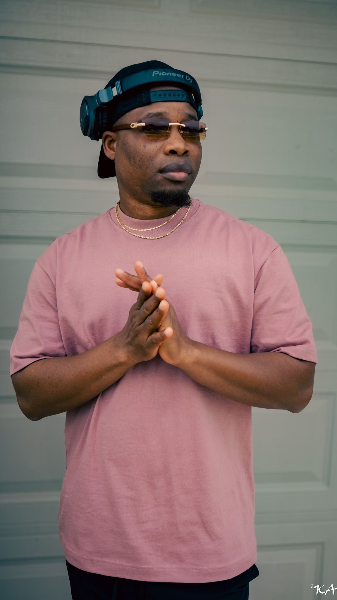Man in pink t-shirt with headphones on cap, gold chain necklace and glasses, hands clasped together outdoors.