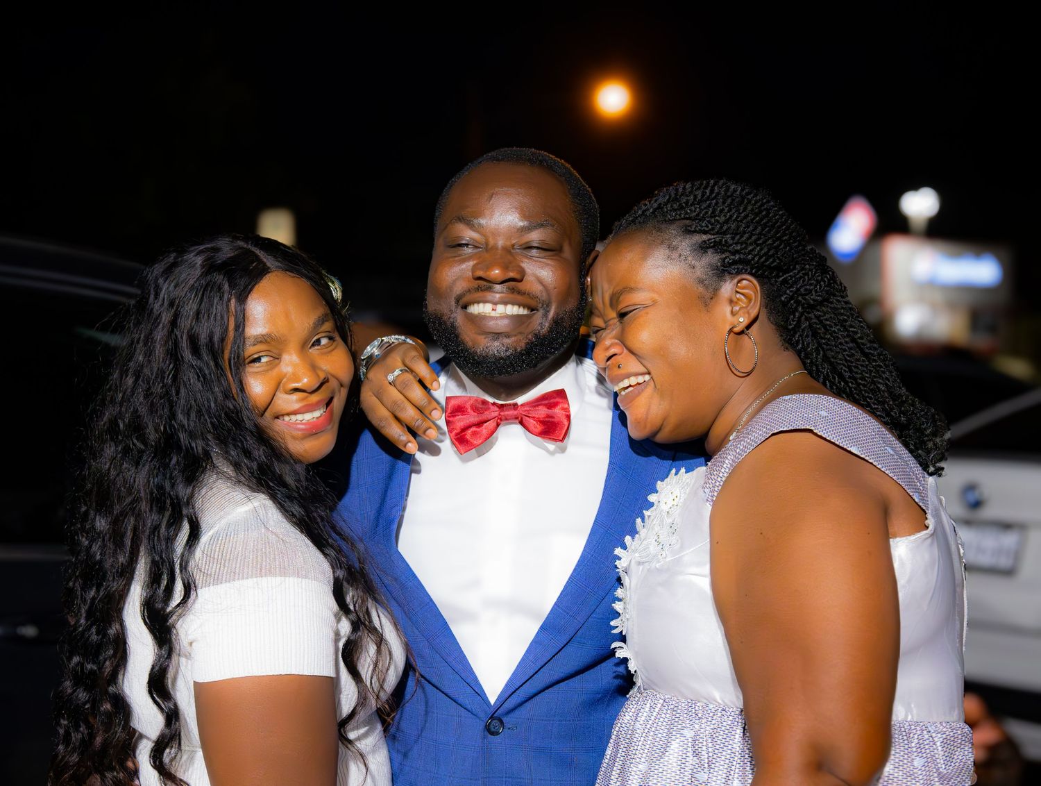 Man in blue suit and red bow tie smiles with two women in white dresses at a formal evening event.