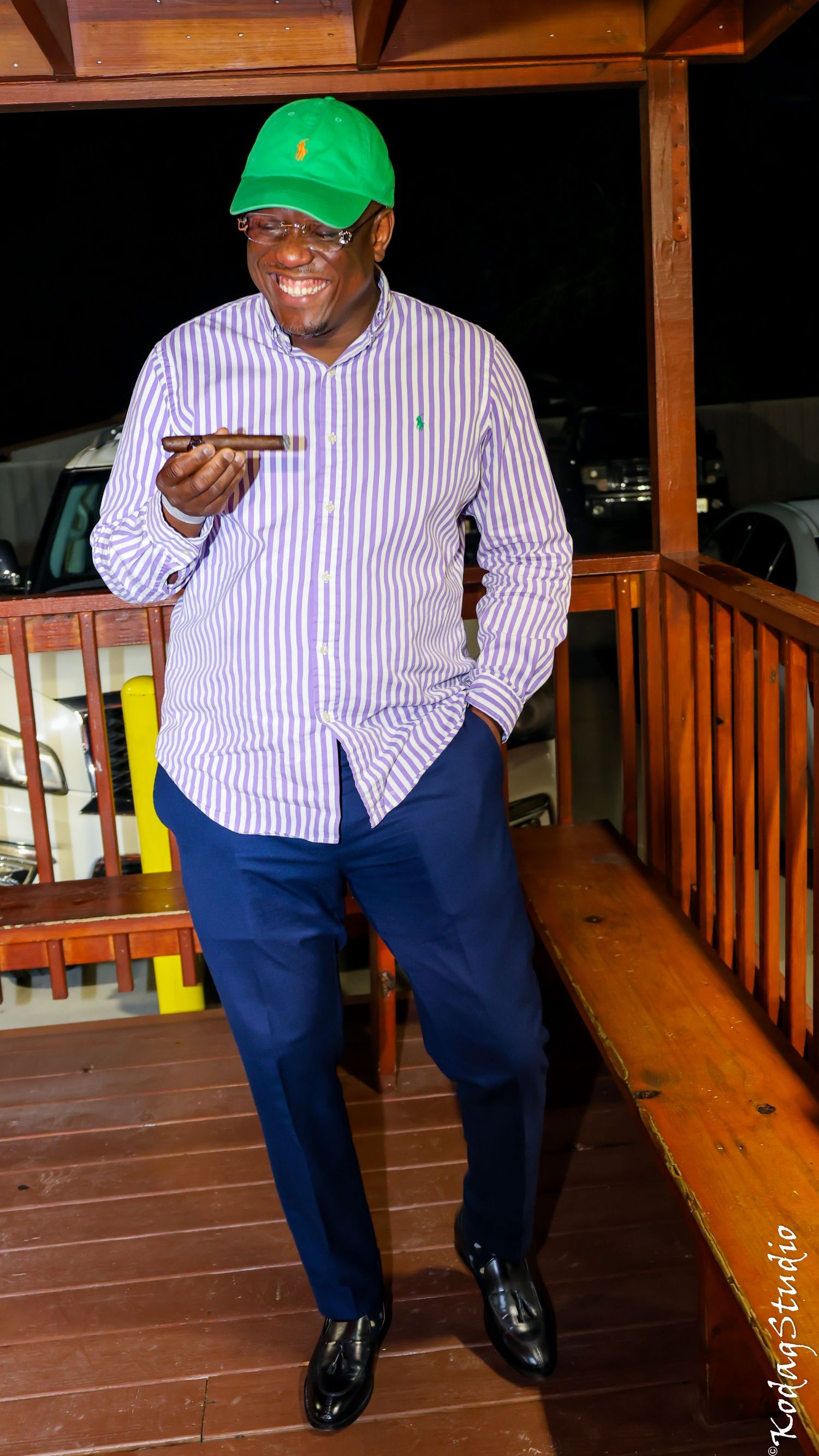 Smiling man in green Ralph Lauren cap, striped shirt, and blue pants standing on wooden deck holding a phone.