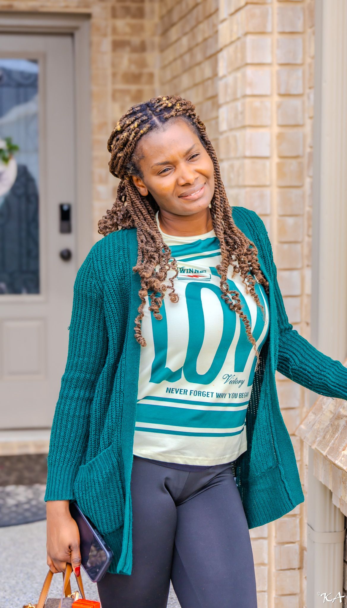 Woman with braided hair wearing a teal cardigan over a graphic tee and leggings, smiling outdoors near a brick building.