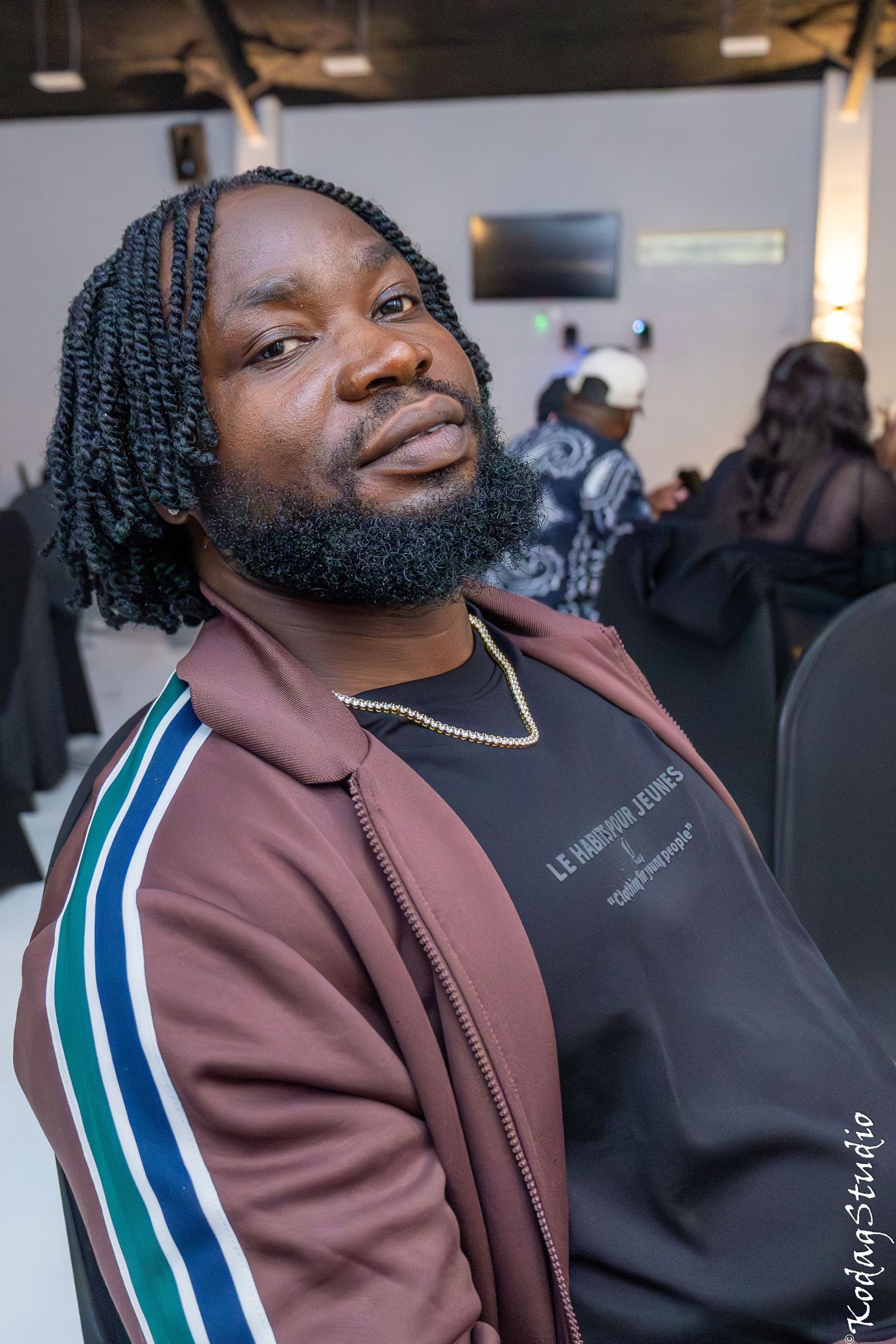 A bearded Black man with braided hair wearing a maroon jacket and gold chain poses at an indoor event.