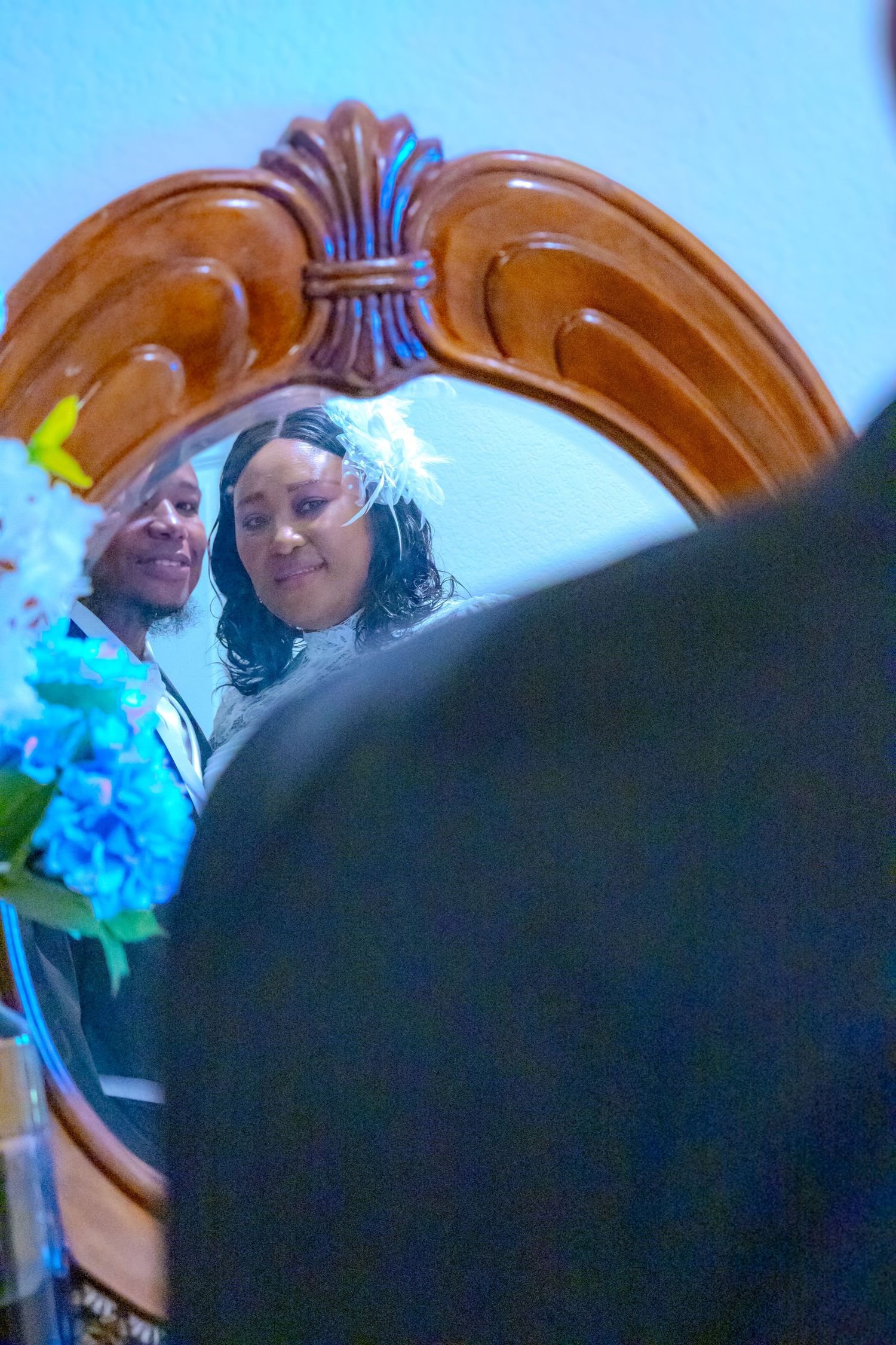 Two women reflected in an ornate wooden mirror, one wearing a white feathered hair accessory at a special event.