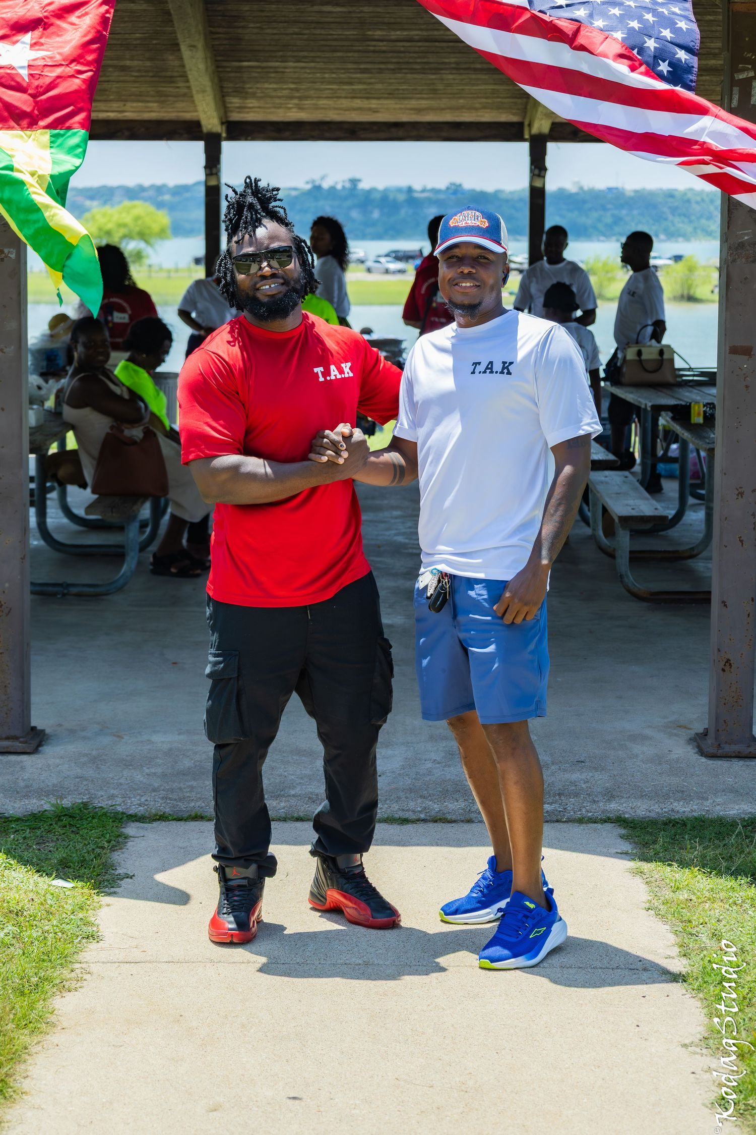 Two men wearing matching T.A.K. shirts shake hands under a pavilion near a lake with an American flag overhead.