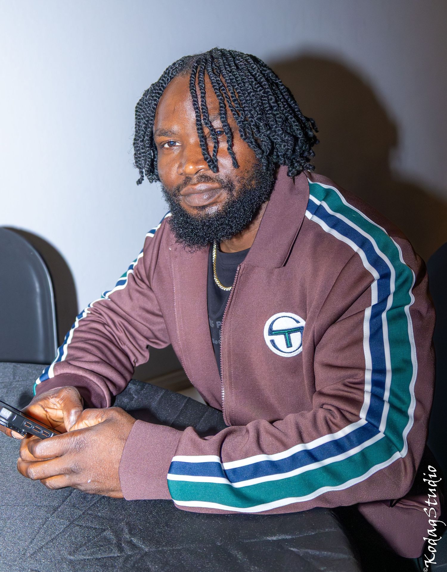 Bearded man with braids wearing a maroon Sergio Tacchini track jacket, holding a phone at an indoor event.
