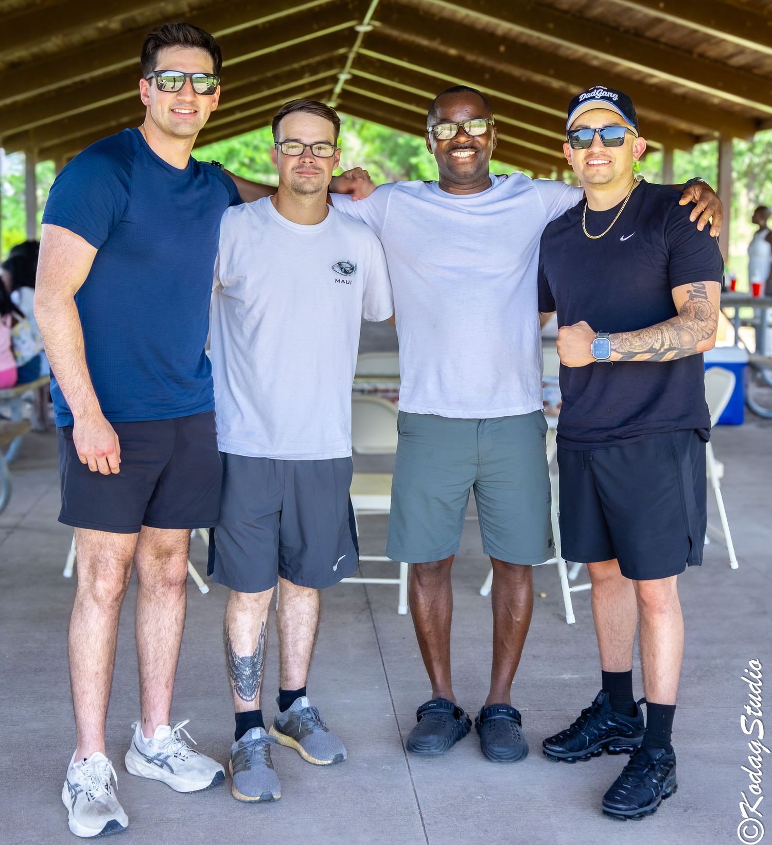 Four men smiling and posing together under a pavilion outdoors on a sunny day.