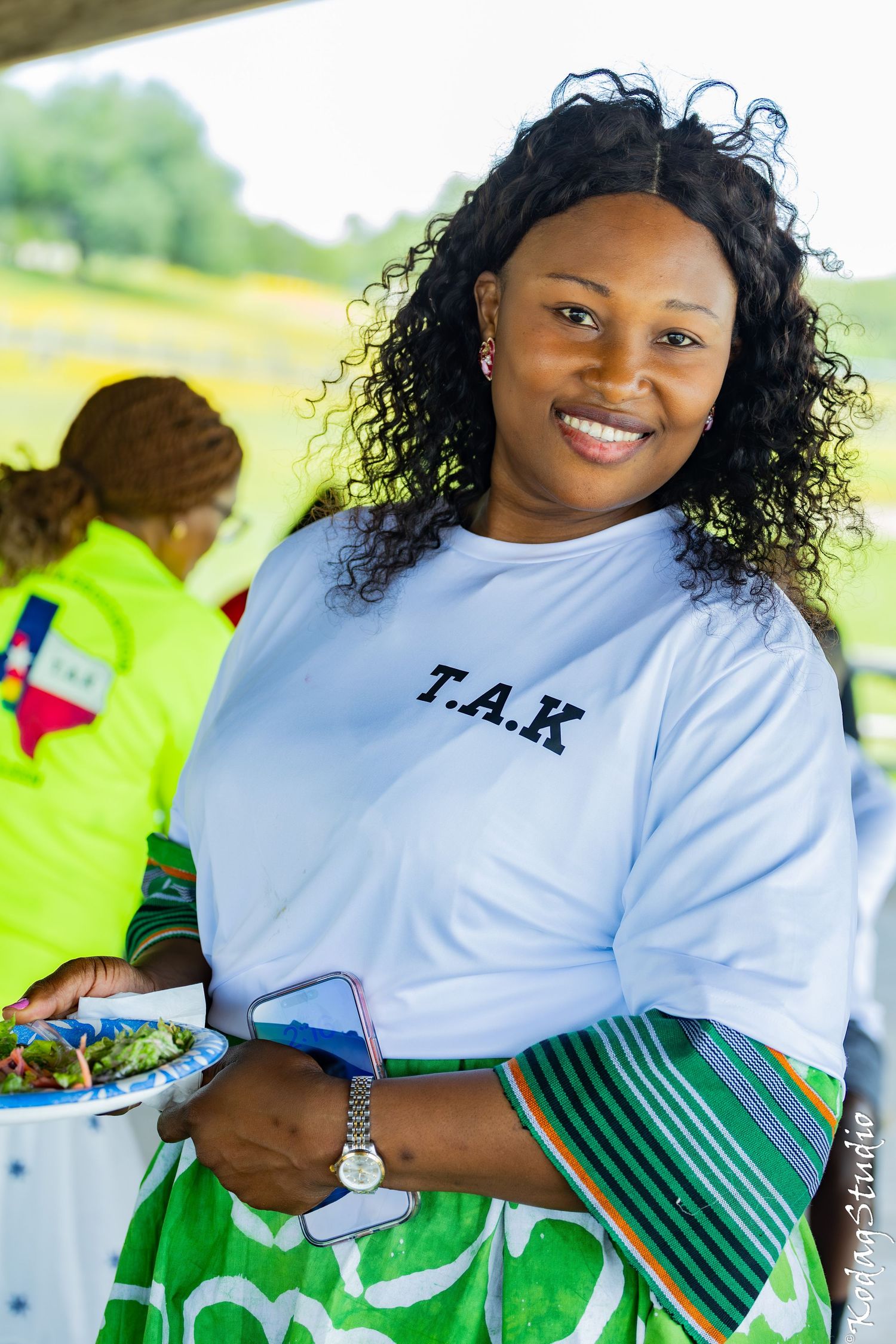Smiling woman in a white T.A.K shirt holding a plate of food and phone at an outdoor event.