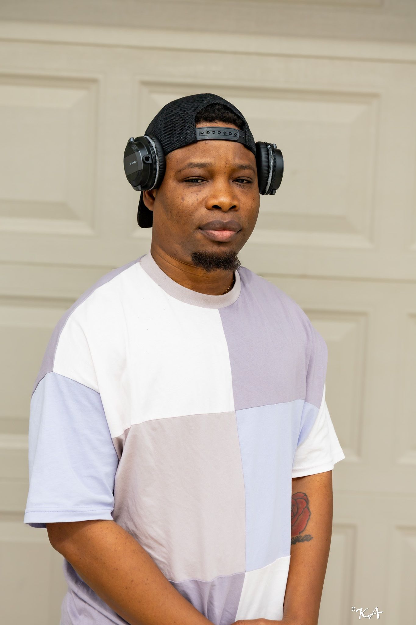Man wearing headphones and snapback cap poses in colorful patchwork t-shirt outdoors.