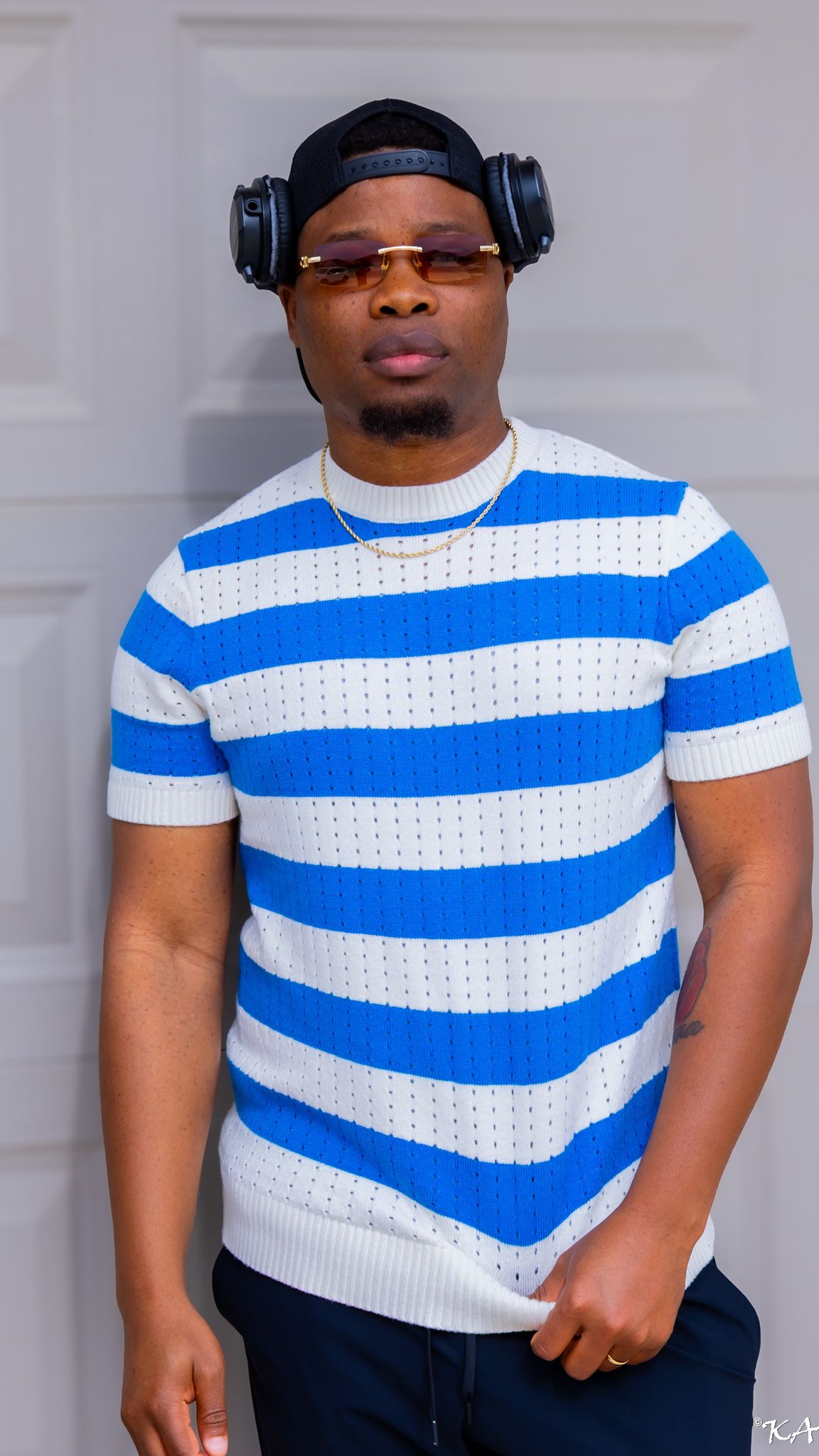 Young man in blue and white striped knit shirt wearing headphones and sunglasses posing confidently indoors.